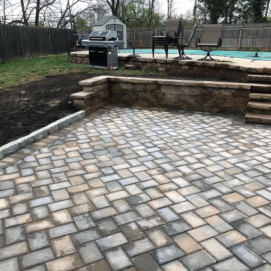 A backyard patio with interlocking stone pavers, a short retaining wall, and steps leading up to a higher level with patio furniture, a grill, and a small shed in the background.