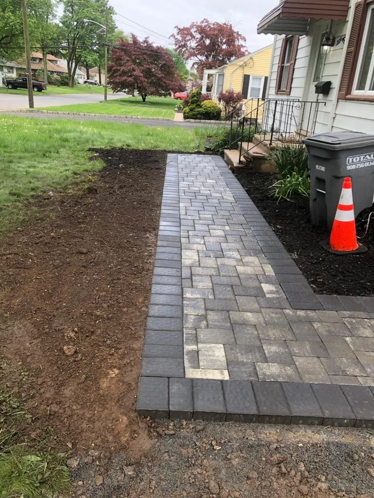 Finished brick pathway leading from the yard to the front steps of a house, with some freshly tilled soil on the side, a trash bin, and a traffic cone nearby.