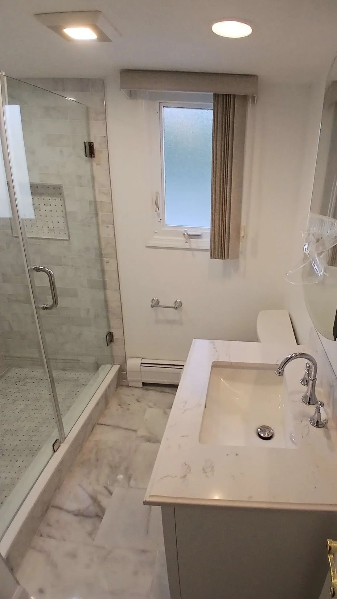 Small bathroom with marble floor, a glass-enclosed shower, a window with beige curtains, and a white marble countertop with a built-in sink and silver faucet.