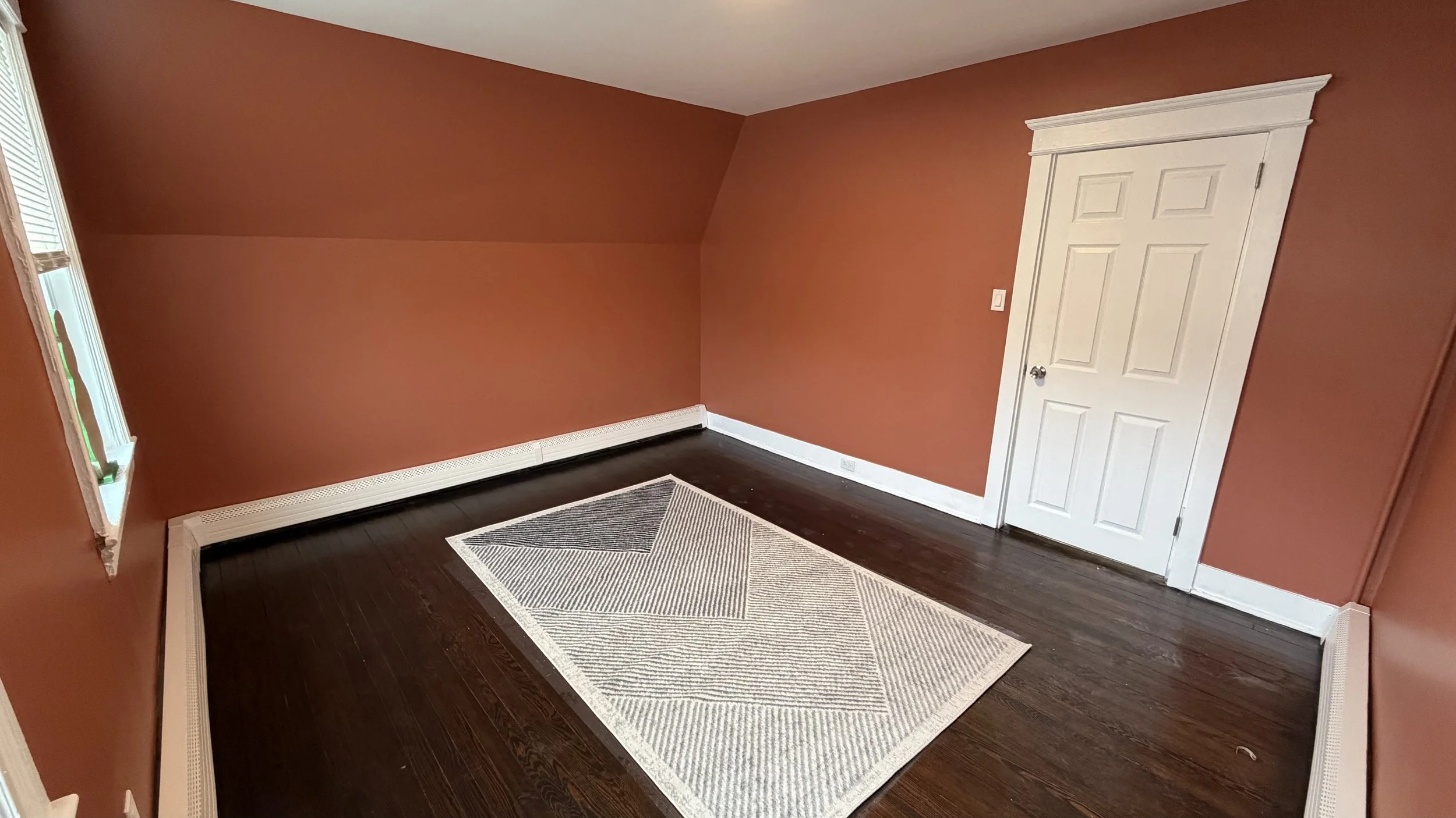 Empty room with brick walls, dark hardwood flooring, a small window, white door, and a geometric patterned rug.