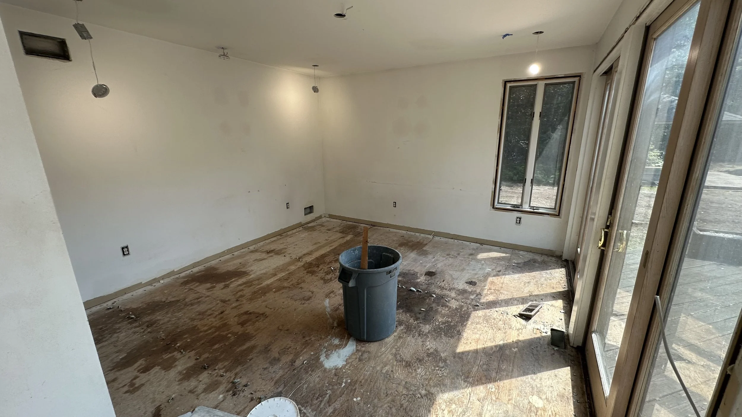 Empty room during renovation with unfinished flooring, white walls, multiple electrical outlets, a window, a door, and some construction tools and debris.