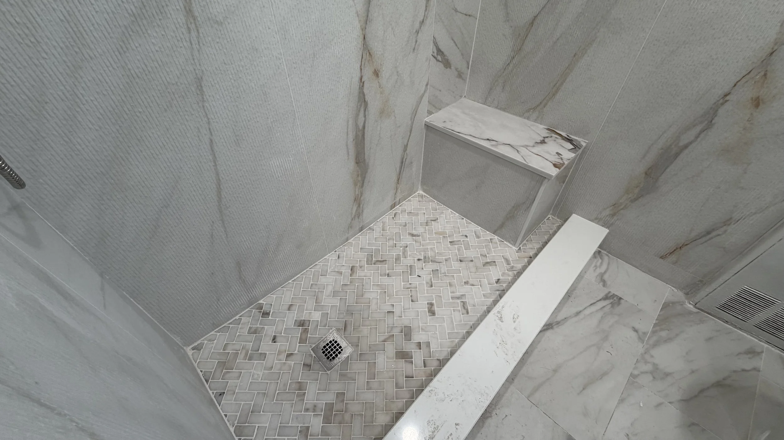 Bathroom shower area with gray tiled walls and small mosaic tile floor, featuring a marble ledge and chrome drain on the floor.