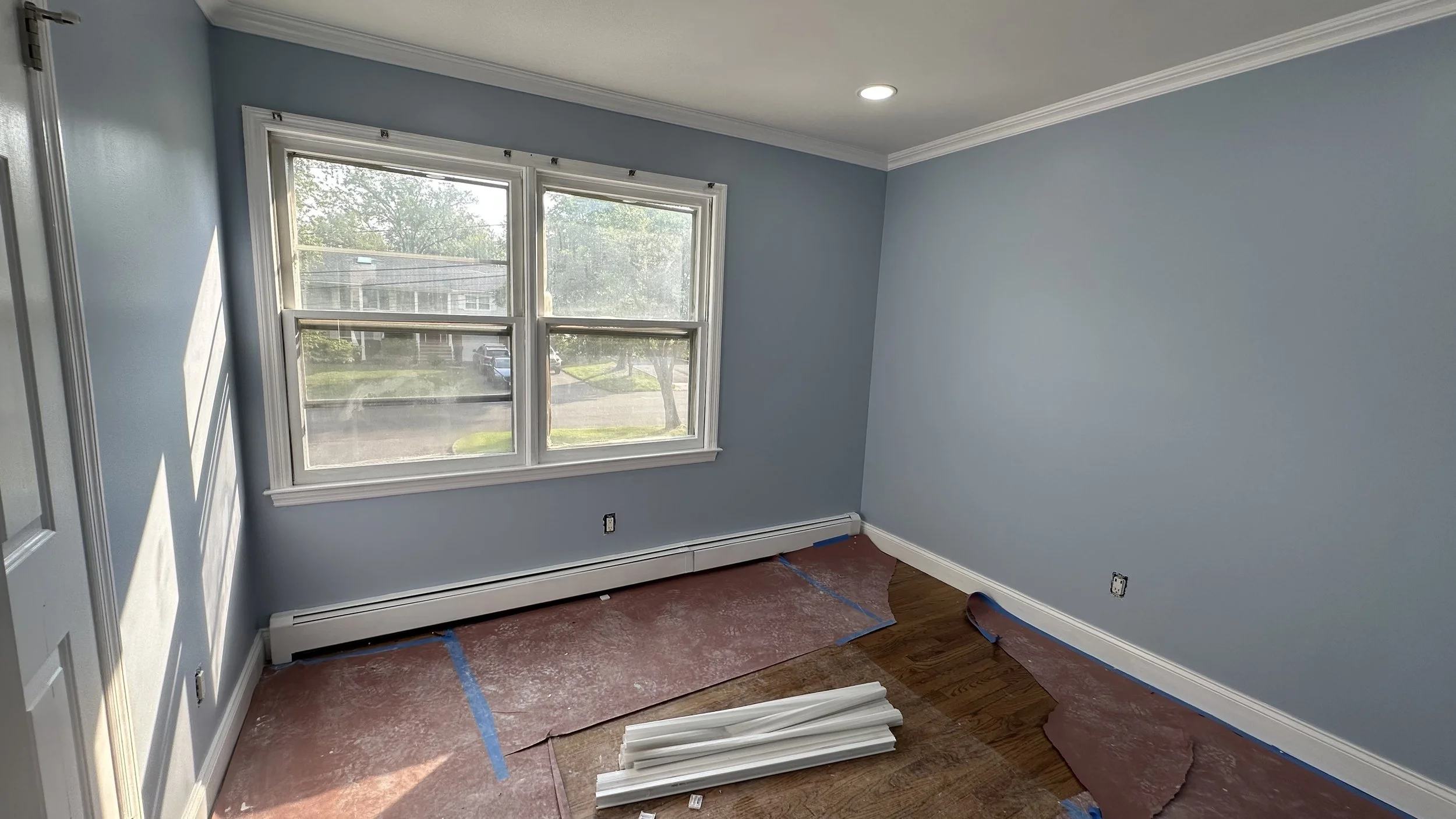 Room with light blue walls, large window, and hardwood floor. The floor is covered with protective pink paper and blue tape, with some white molding placed on the floor. No furniture visible.
