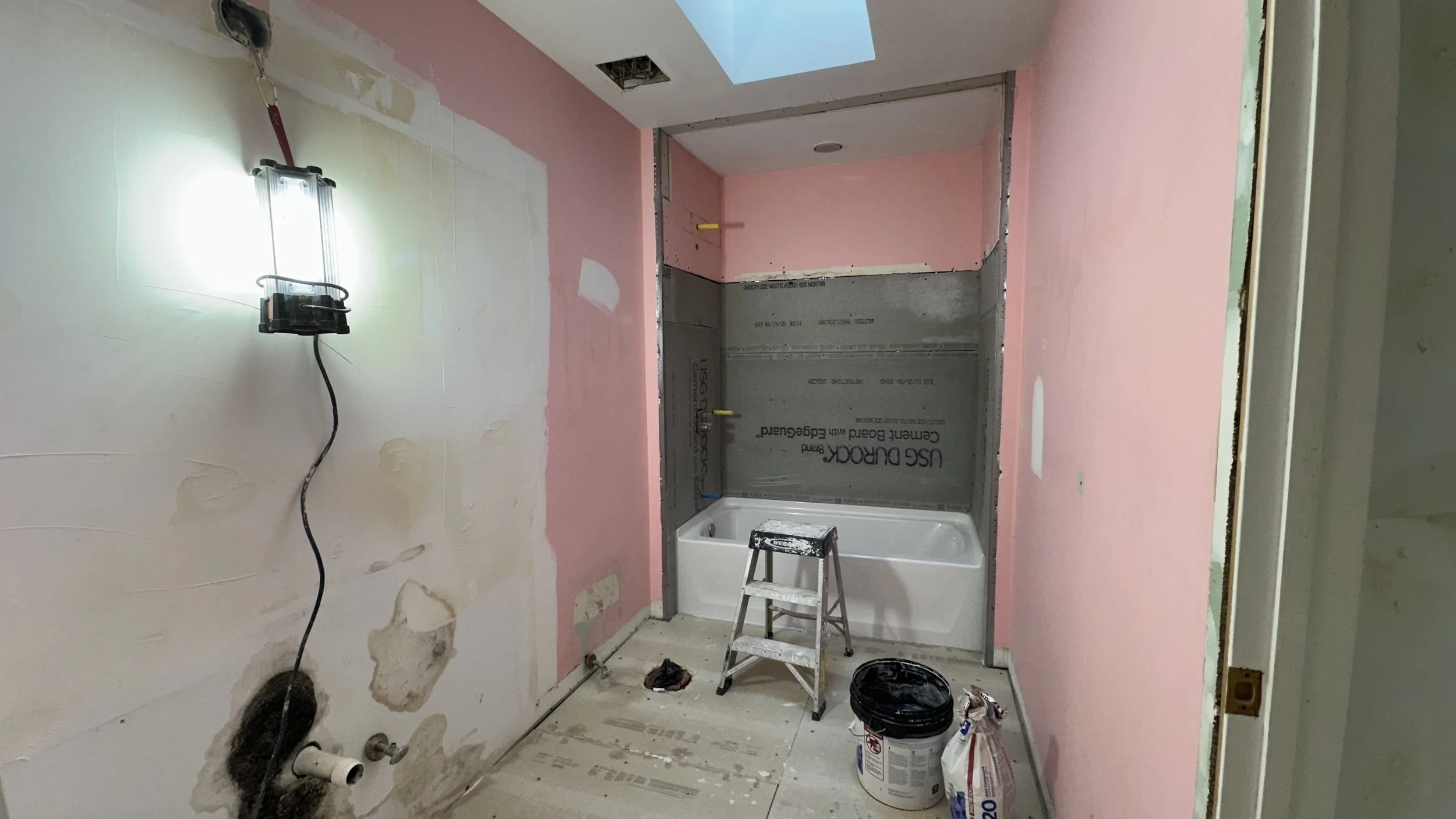 Bathroom under renovation with partially finished pink and white walls, a bathtub, construction tools, a ladder, and ongoing drywall and painting work.