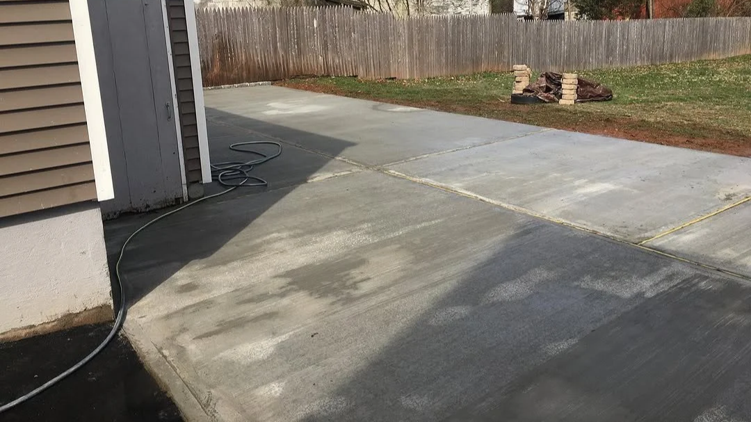 Recently sealed and cleaned concrete driveway in a backyard with a wooden fence and a small pile of bricks in the background.