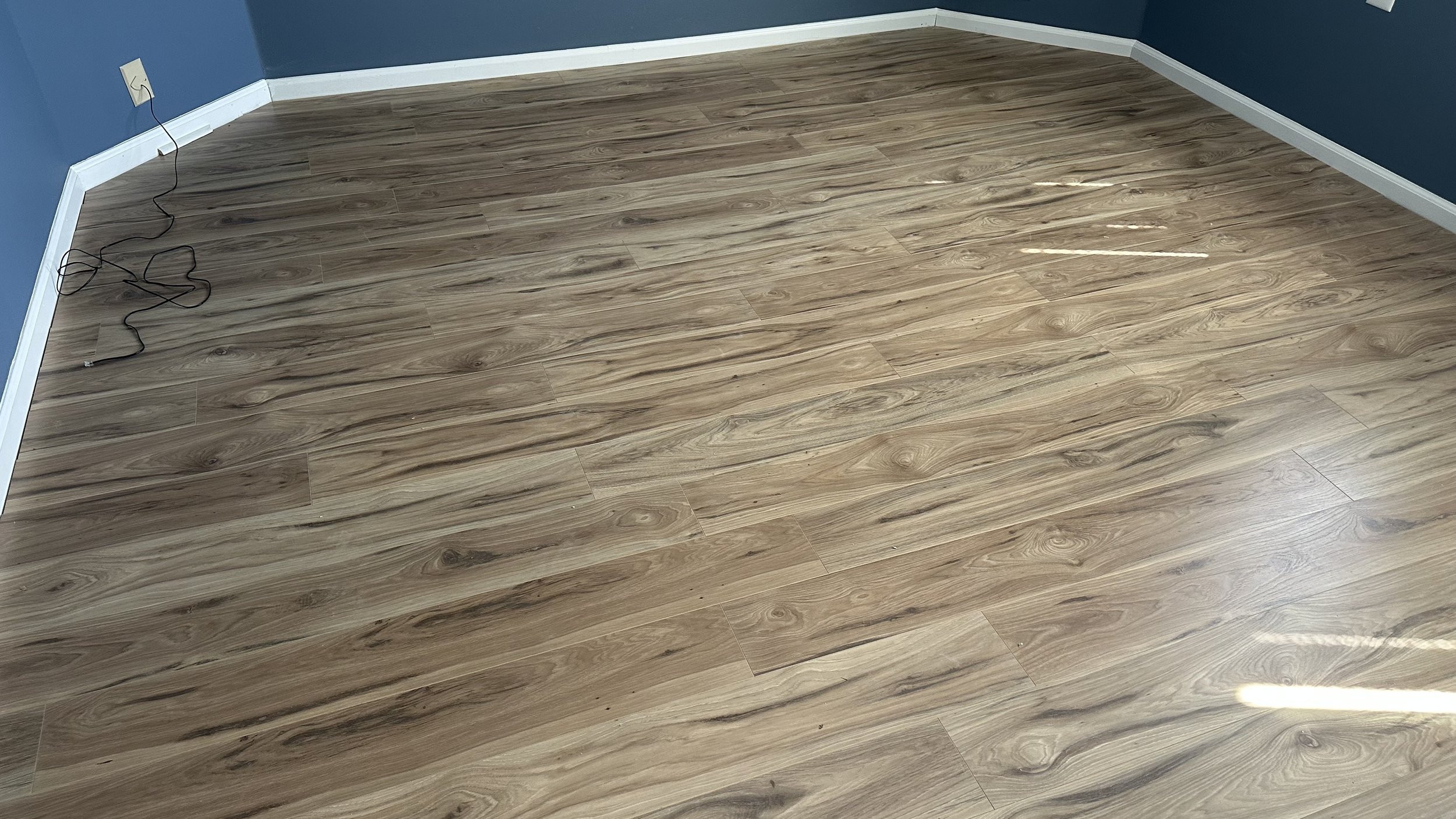 Empty room with wood laminate flooring, blue walls, white baseboards, and an electrical outlet with a black power cord on the floor.