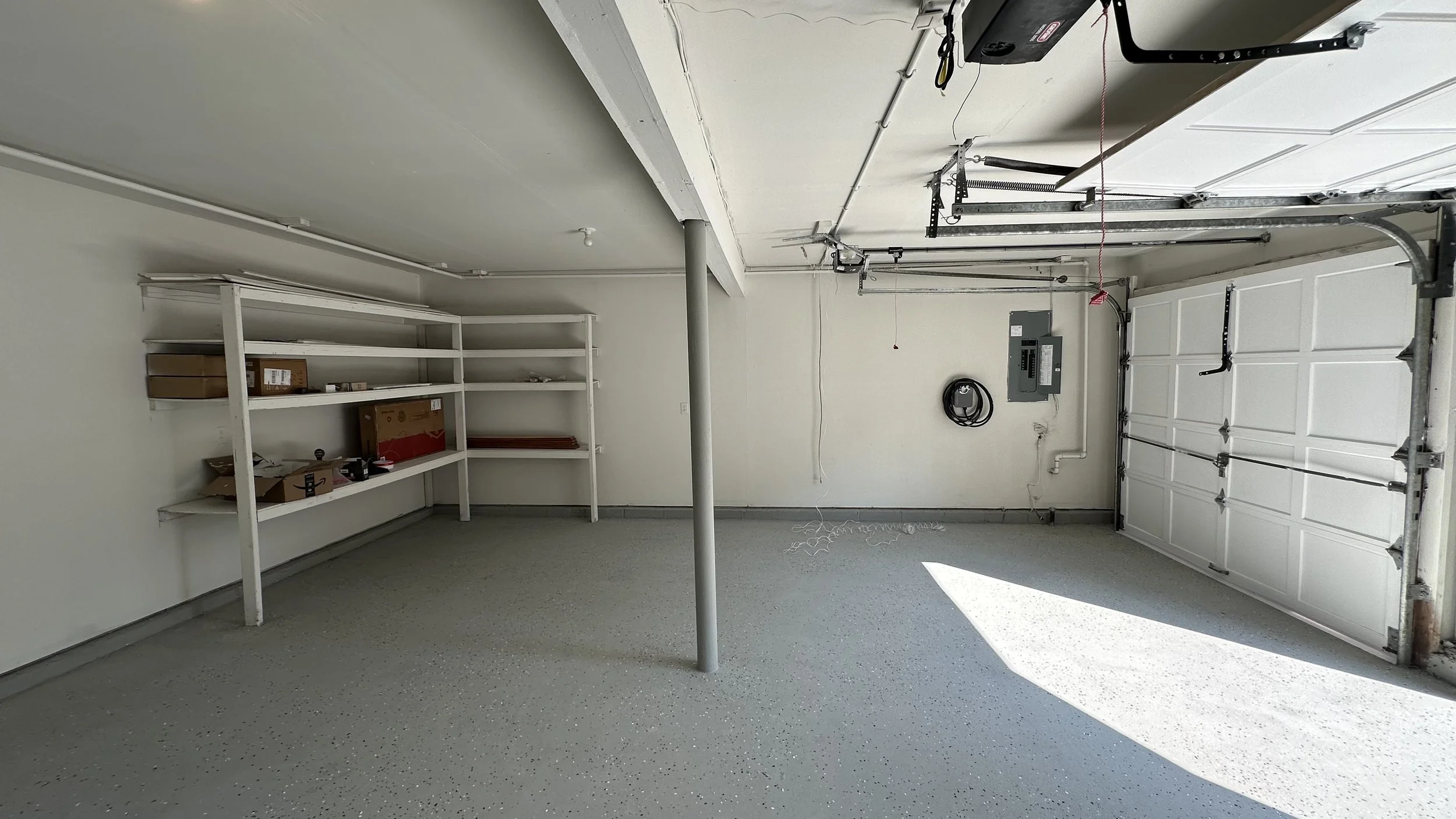 Empty garage with white walls, white shelves on the left, a closed garage door on the right, and a small gray pole in the center. There are electrical panels and a coil of black cable on the wall at the back.