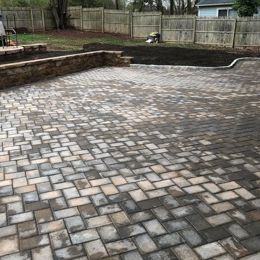 A newly paved backyard with a stone patio, a raised stone planter wall, and a dirt area with landscaping in progress, surrounded by a wooden fence.
