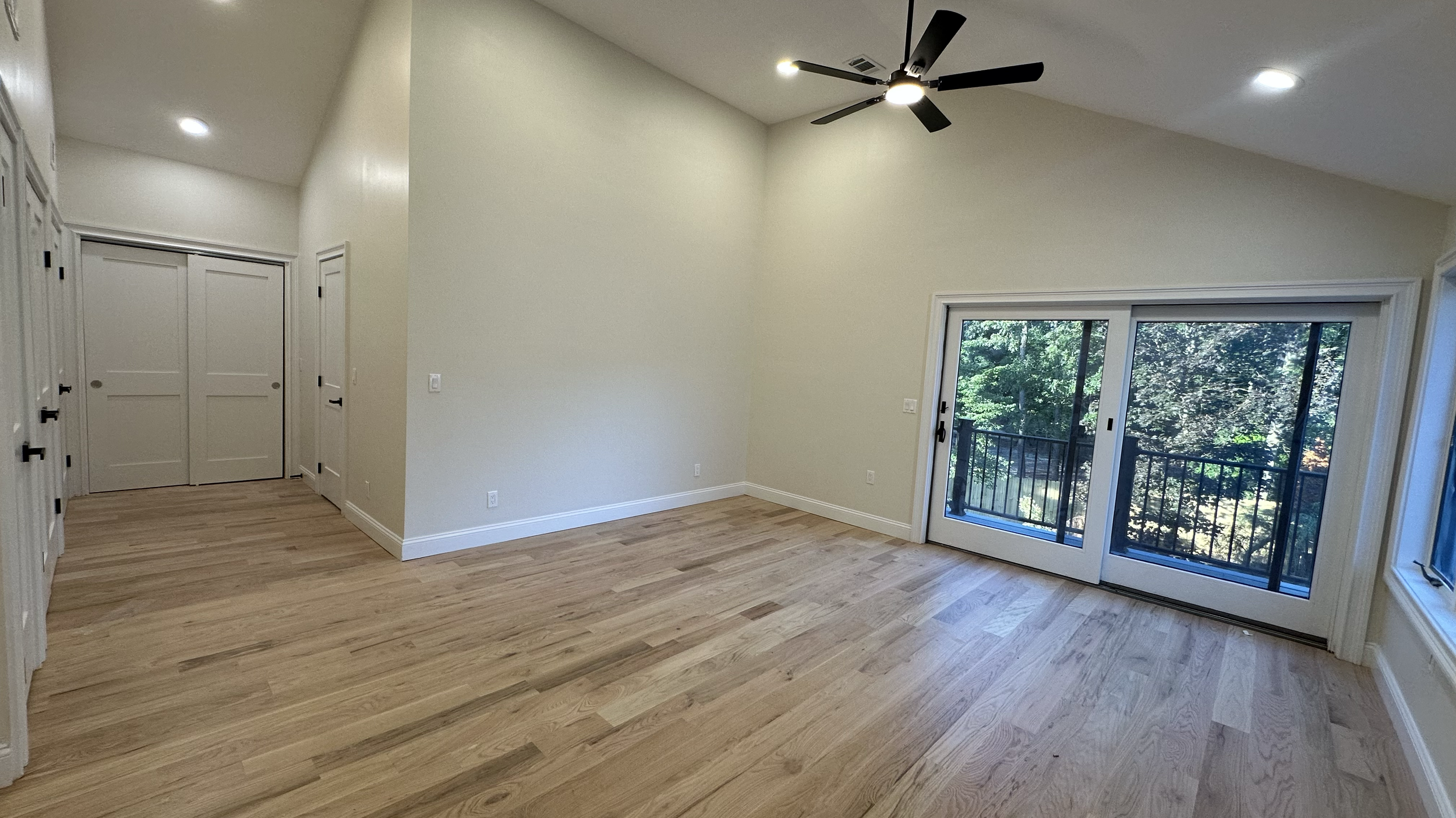Empty room with wood flooring, white walls, large sliding glass door leading to a balcony, and ceiling fan.