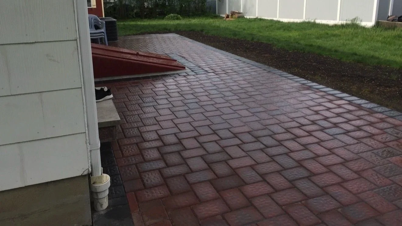 Newly paved red brick patio with a house wall on the left and a grassy yard in the background.