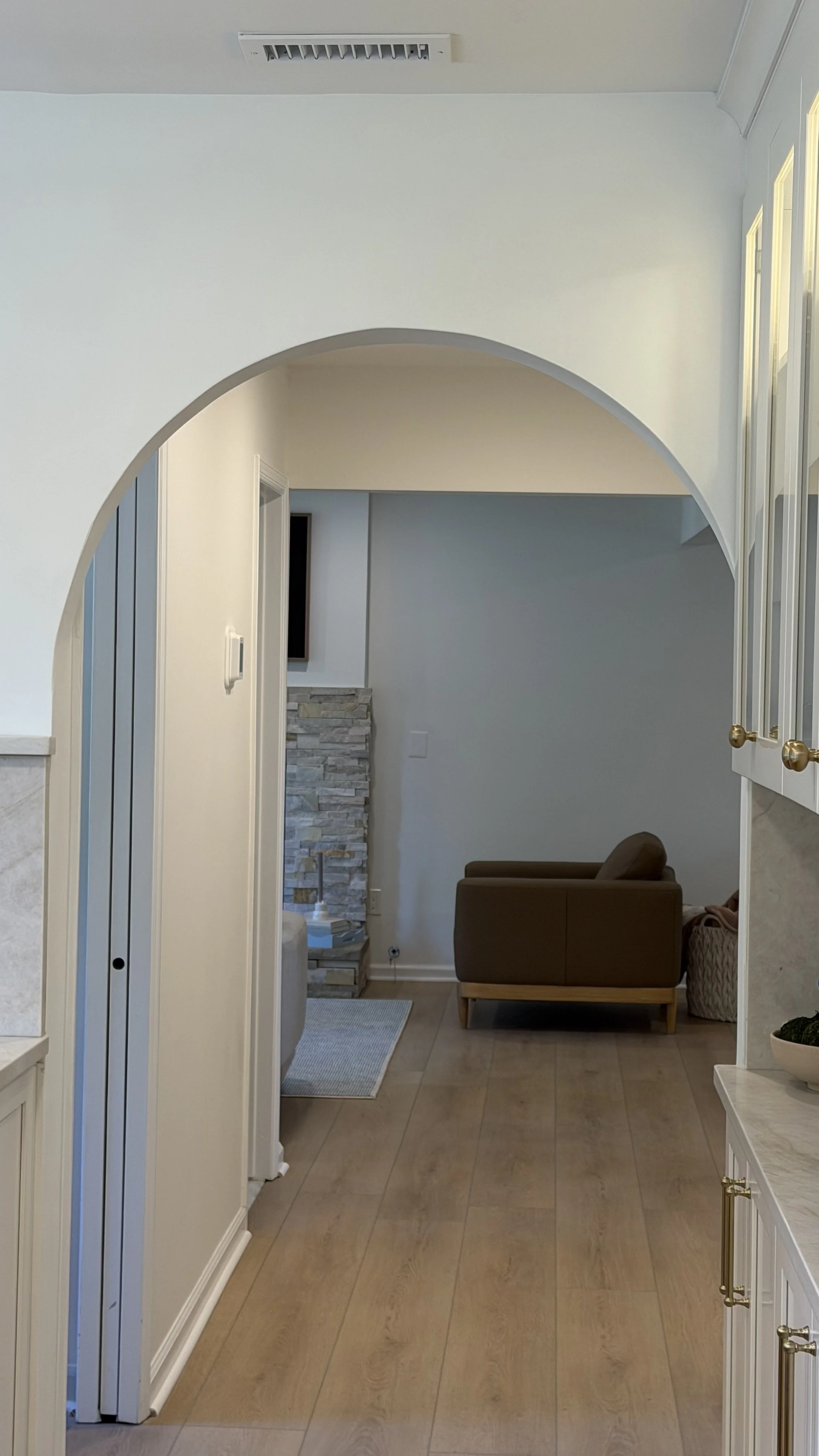 Interior view of a living room through an arched doorway, featuring a brown armchair, a stone fireplace, and a white wall with a mounted television.