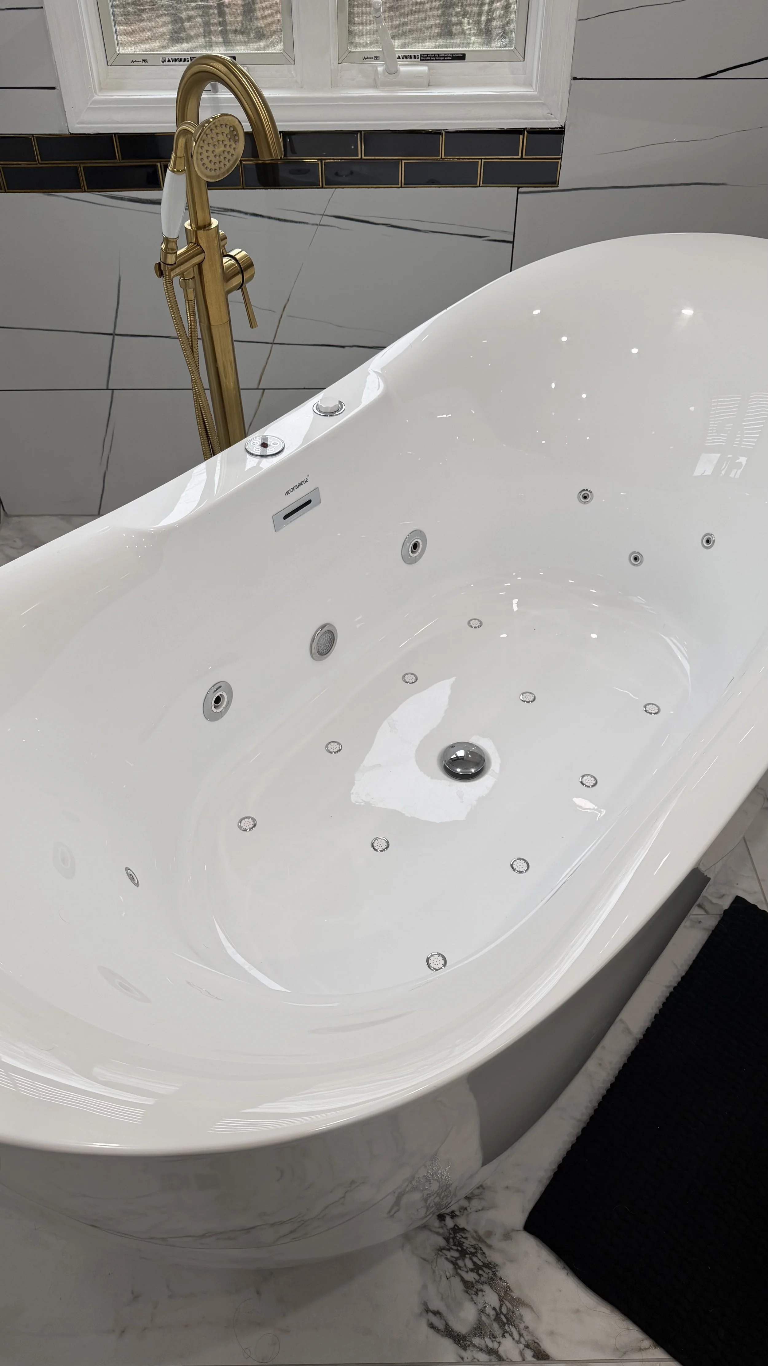 A modern white bathtub with jet nozzles, faucet, and controls, positioned in a bathroom with marble floors and walls, near a window.