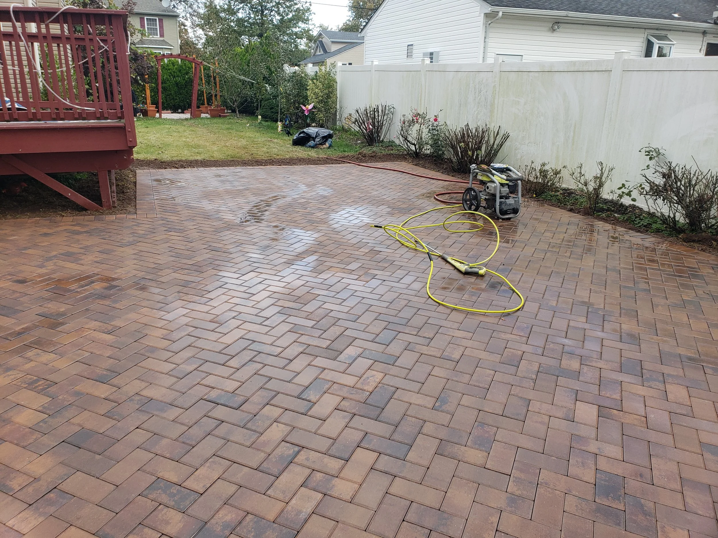 Cleaned brick patio with a power washer, yellow and red hoses, and a white fence in a backyard.