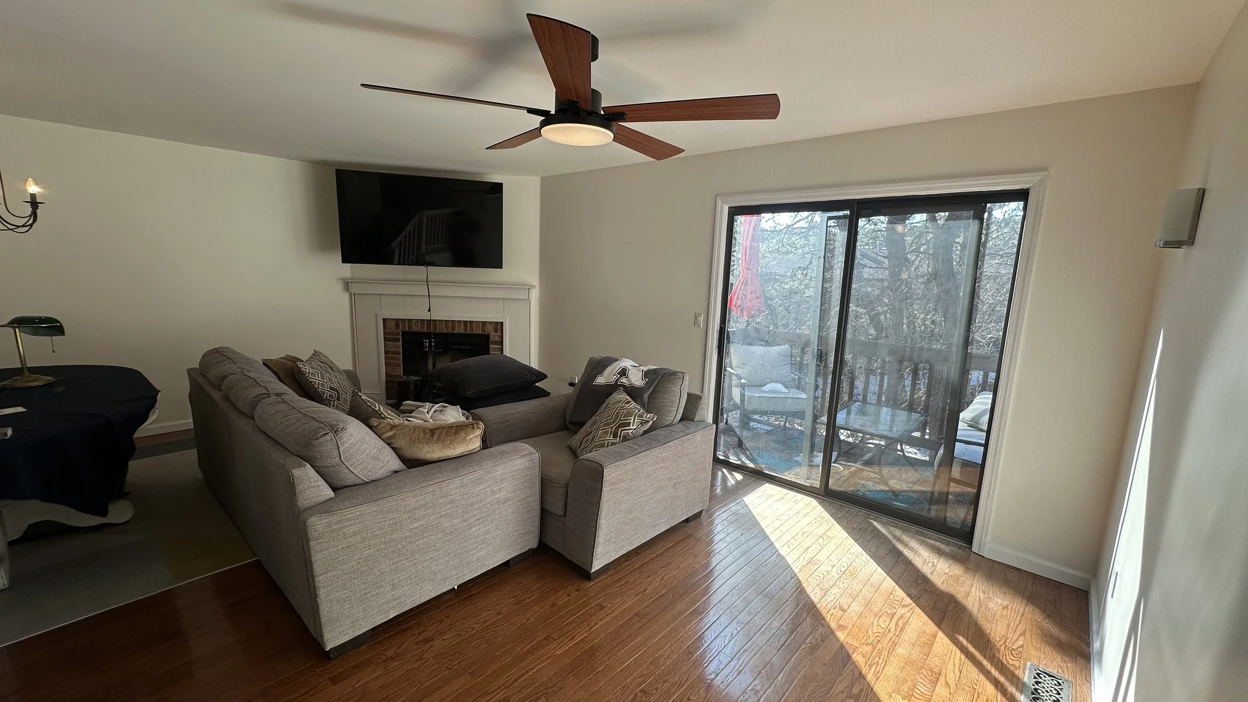 Living room with a gray sectional sofa, a wall-mounted TV above a fireplace, a glass sliding door leading to an outdoor balcony, and wooden flooring.