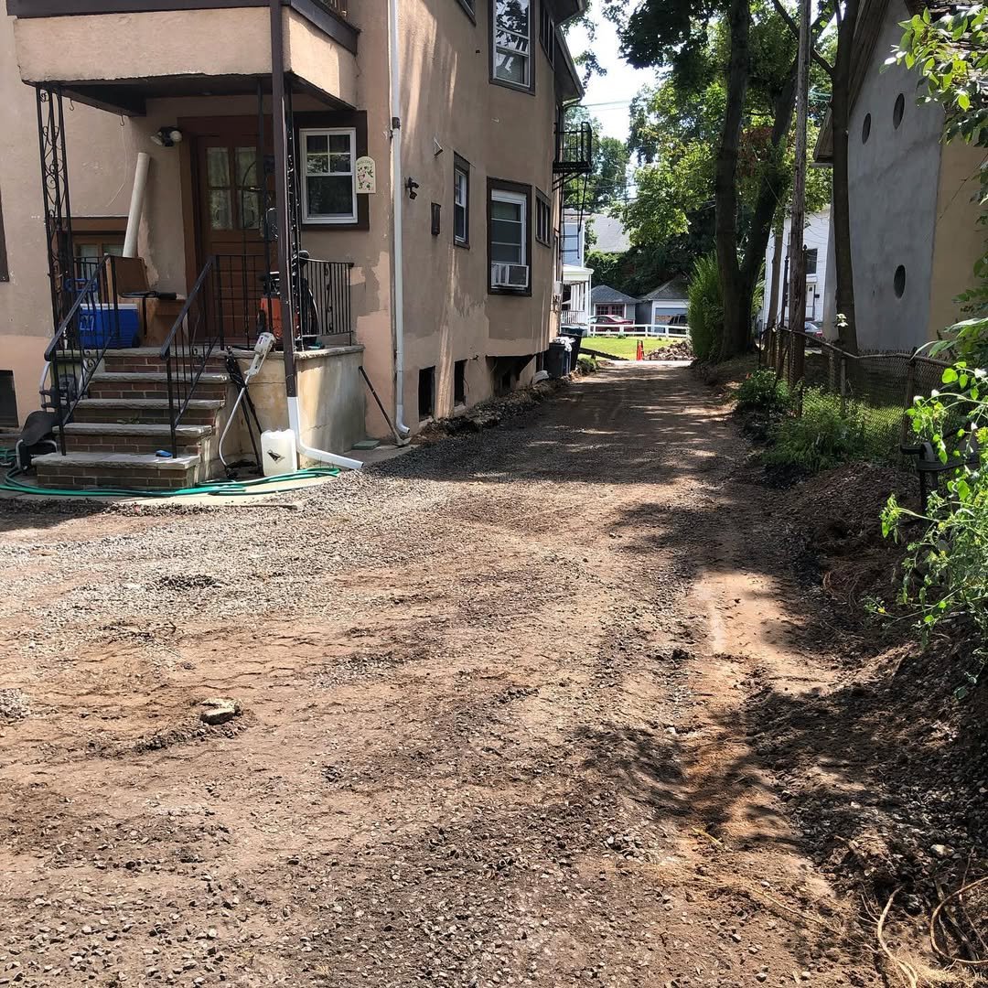A backyard with an unfinished dirt and gravel driveway, bordered by a multi-story beige house with brick stairs and black iron railing on the left, and a fenced garden area with greenery on the right. In the background, there are neighboring houses a