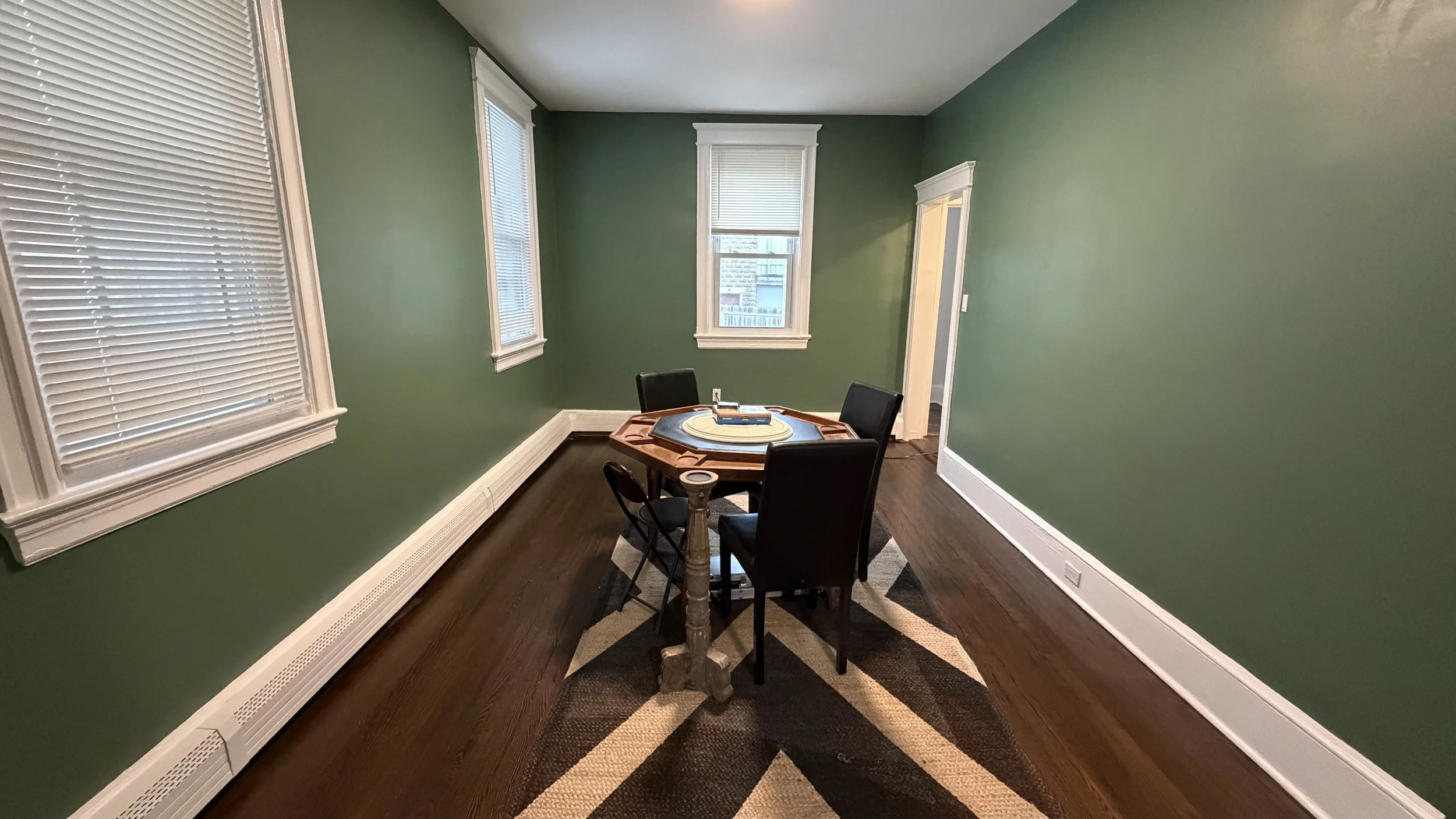 Empty dining room with four windows, green walls, dark wooden floor, and a round table with four chairs.