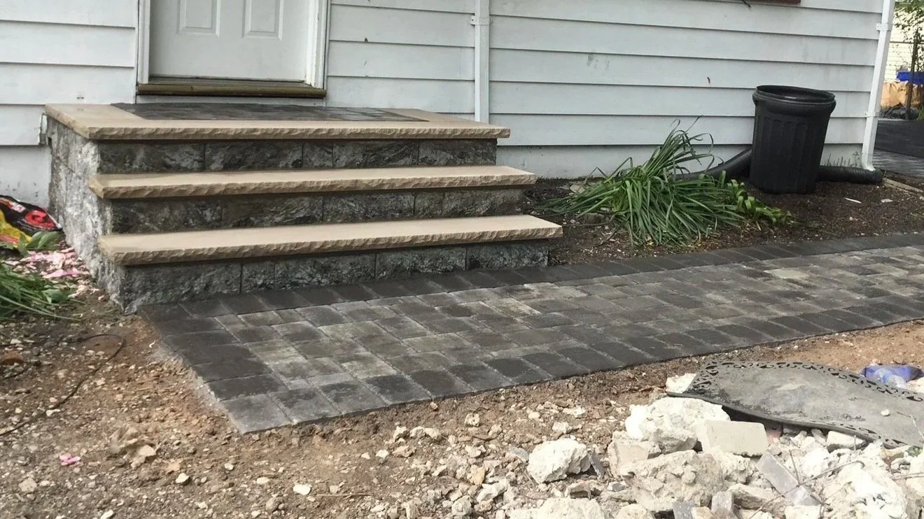 Construction site with a small set of stone steps leading to a door, a paved walkway, a black trash can, some green plants, and loose gravel and dirt in the foreground.