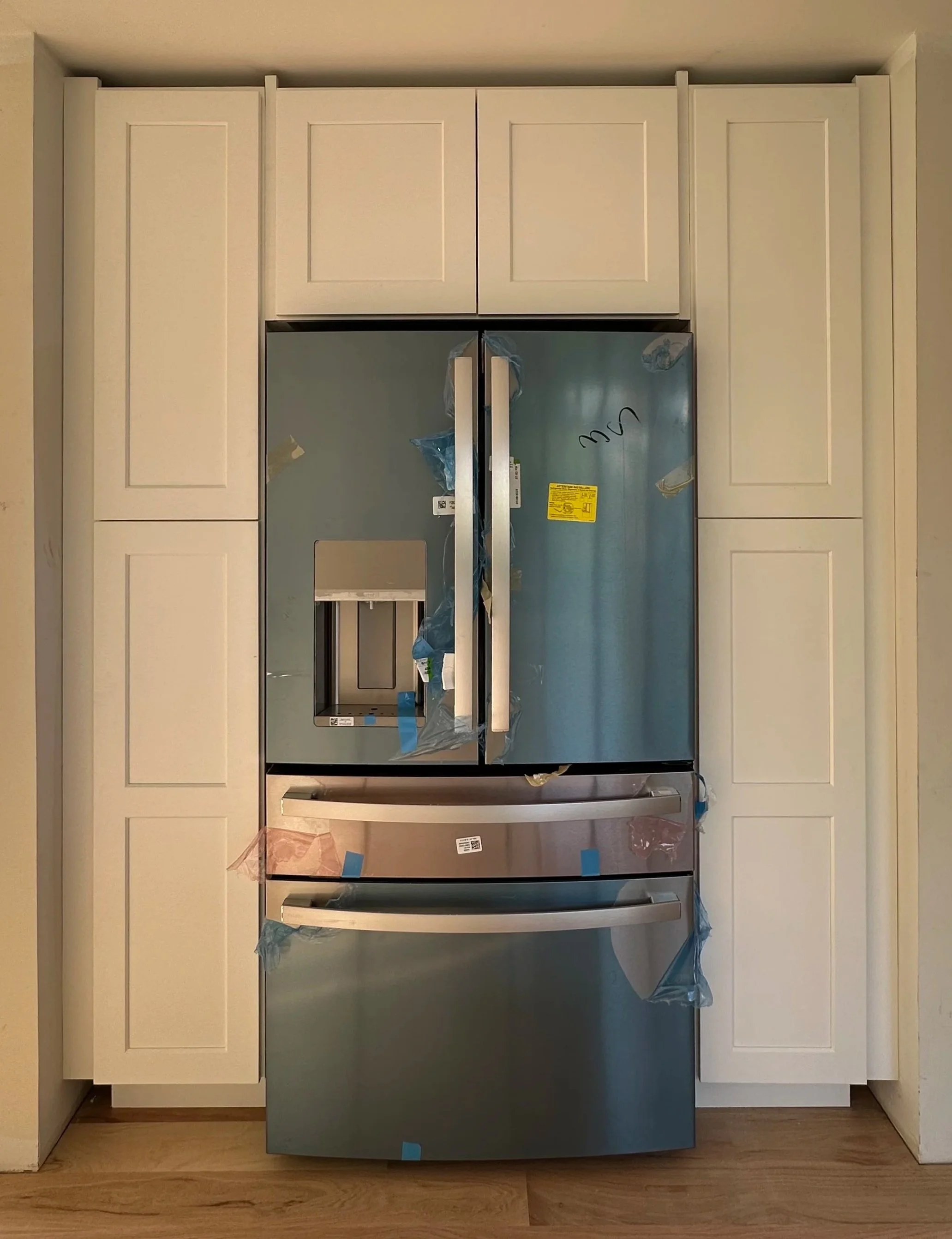 New stainless steel refrigerator with double doors surrounded by white cabinetry in a kitchen.