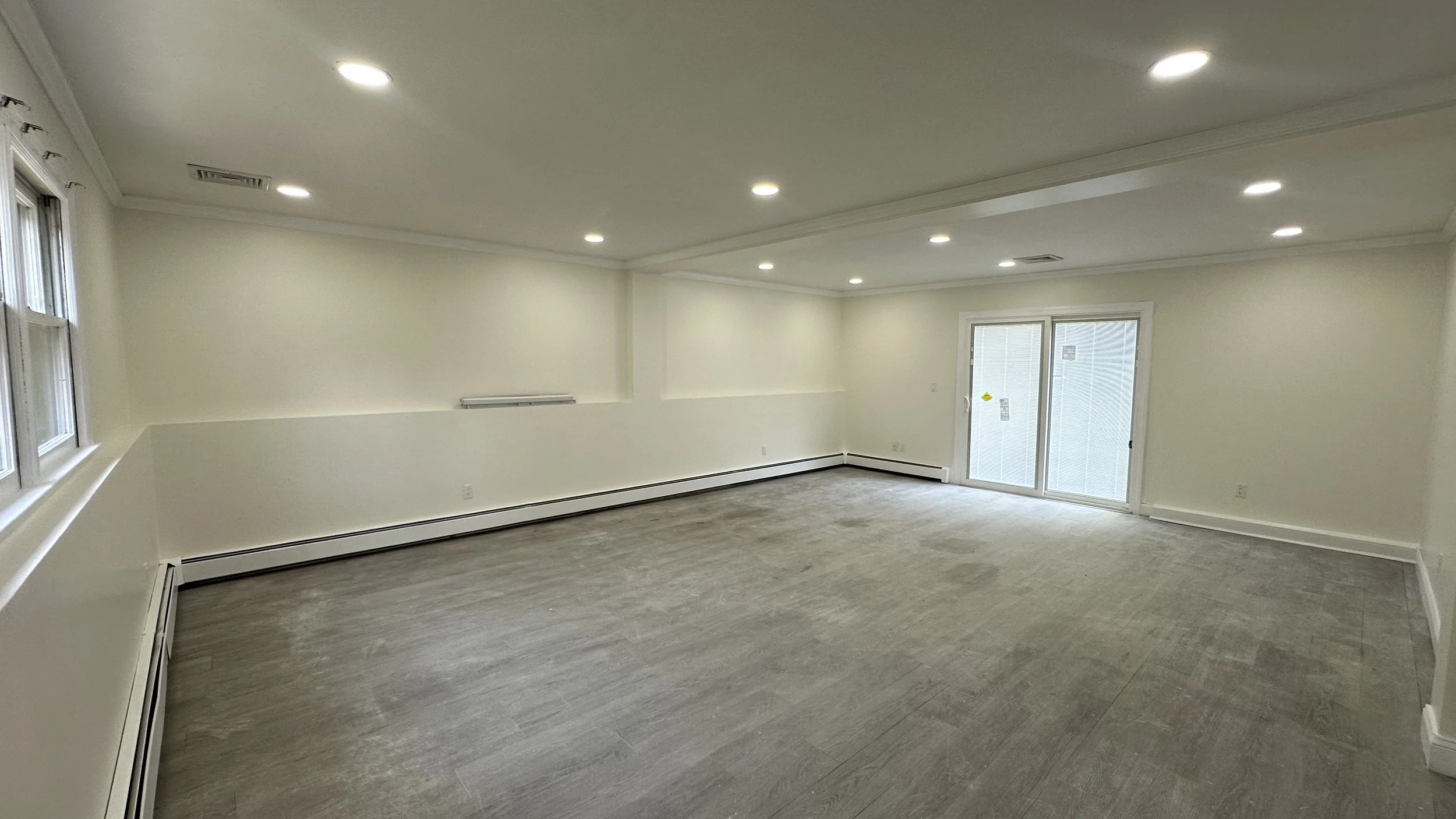 Empty room with gray wood flooring, white walls, ceiling with recessed lights, windows on the left, and sliding glass door on the far wall.