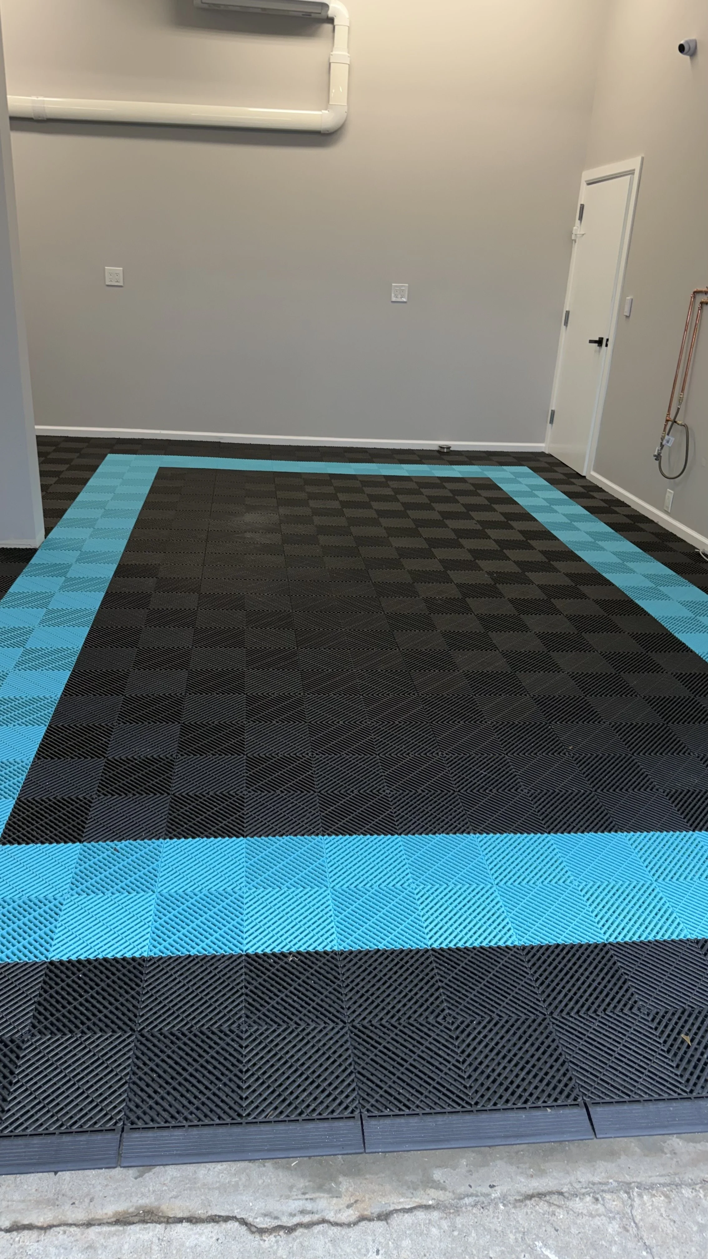 The image shows a section of a room with a checkered black and gray rubber floor mat with a blue border. There are electrical outlets on the wall, a door on the right, and some piping along the upper part of the wall. The flooring appears to be in a garage or utility room.