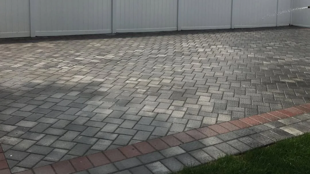 A paved driveway with interlocking gray pavers, bordered by a row of red bricks, next to a small patch of green grass and a gray metal fence in the background.
