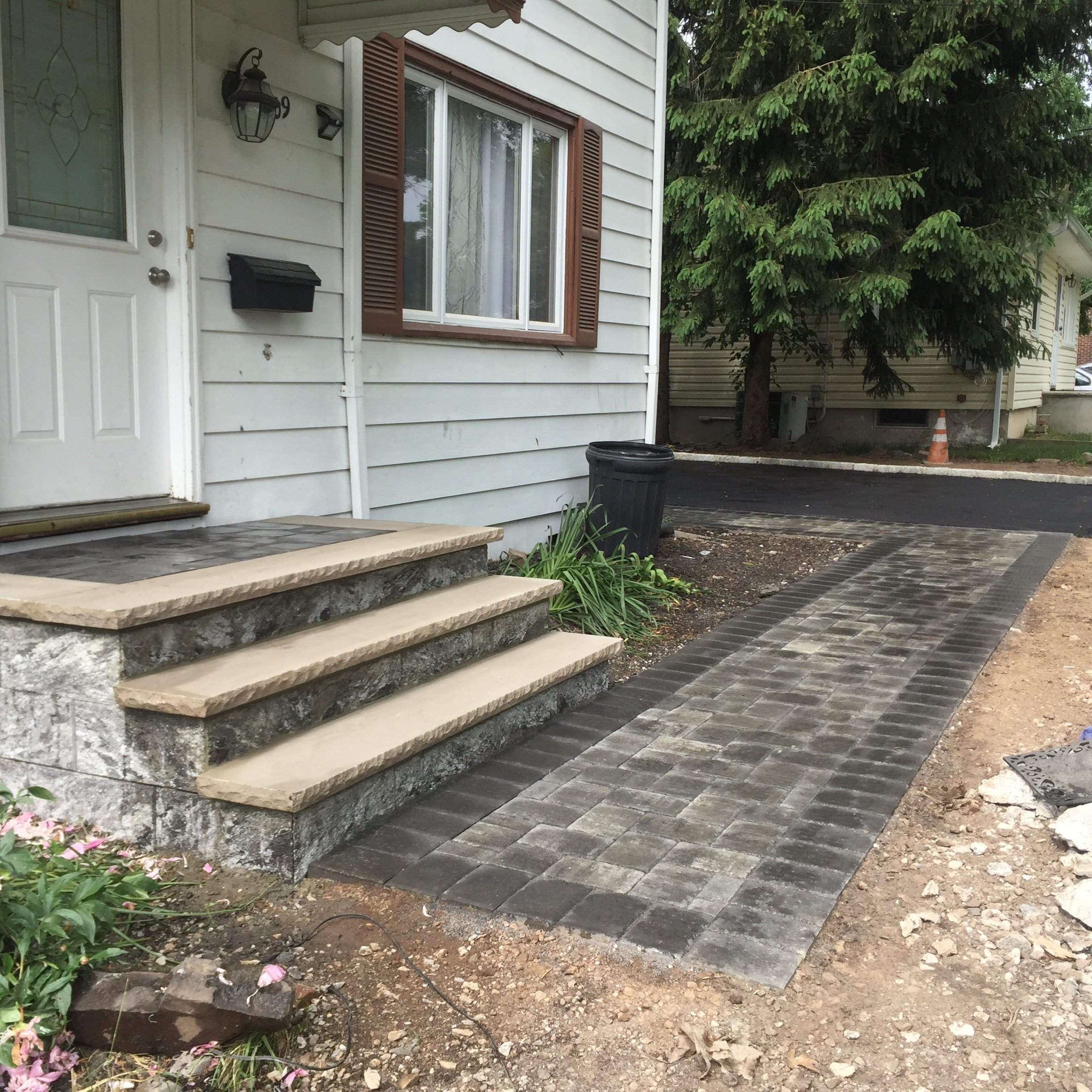 Front steps and walkway of a house with white siding, brown shutters, and a black mailbox, nearby a black trash can, with trees and a street in the background.