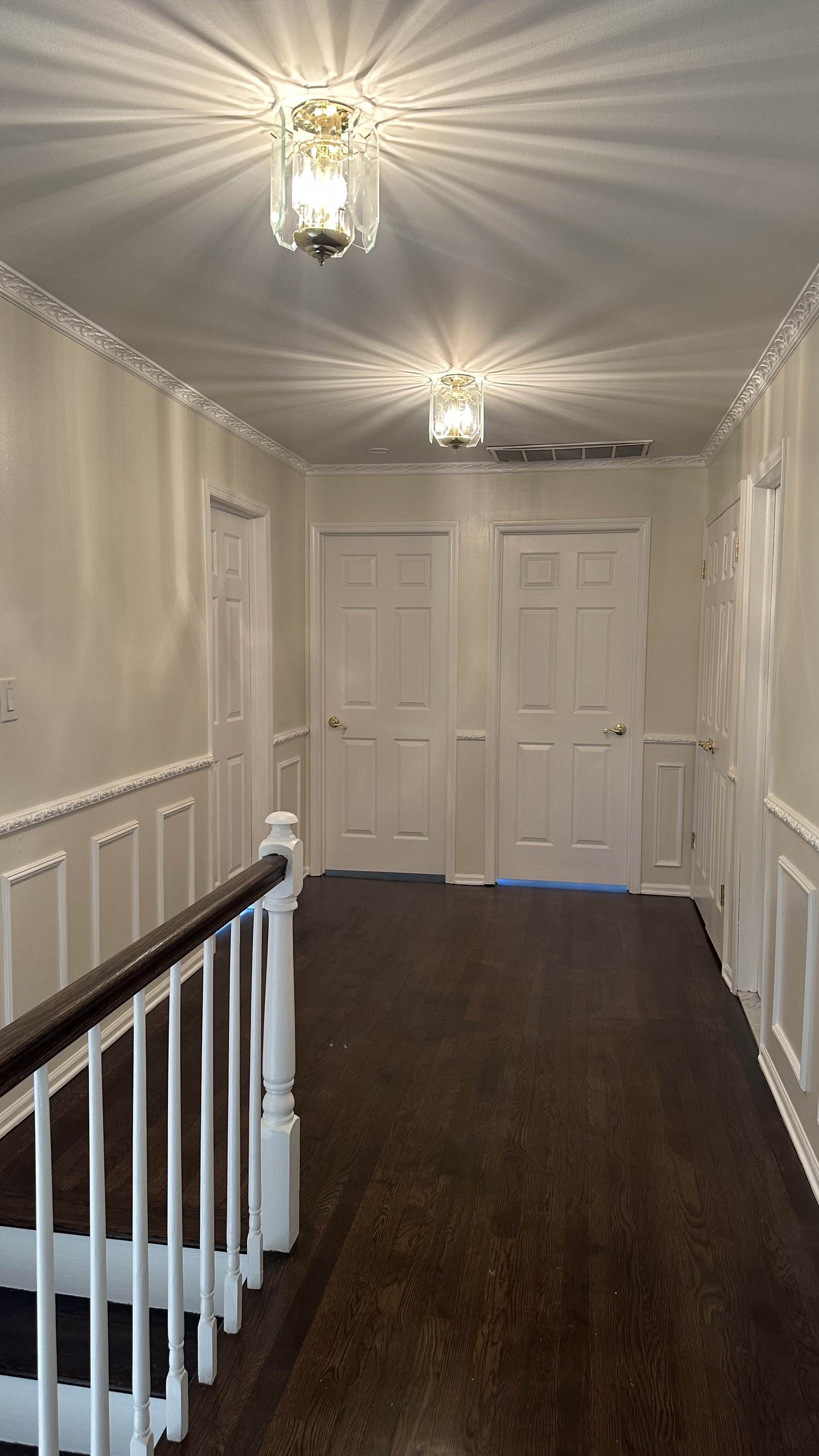 Empty hallway with dark wooden floor, cream walls with decorative molding, four closed white doors, and two ceiling light fixtures casting bright light.