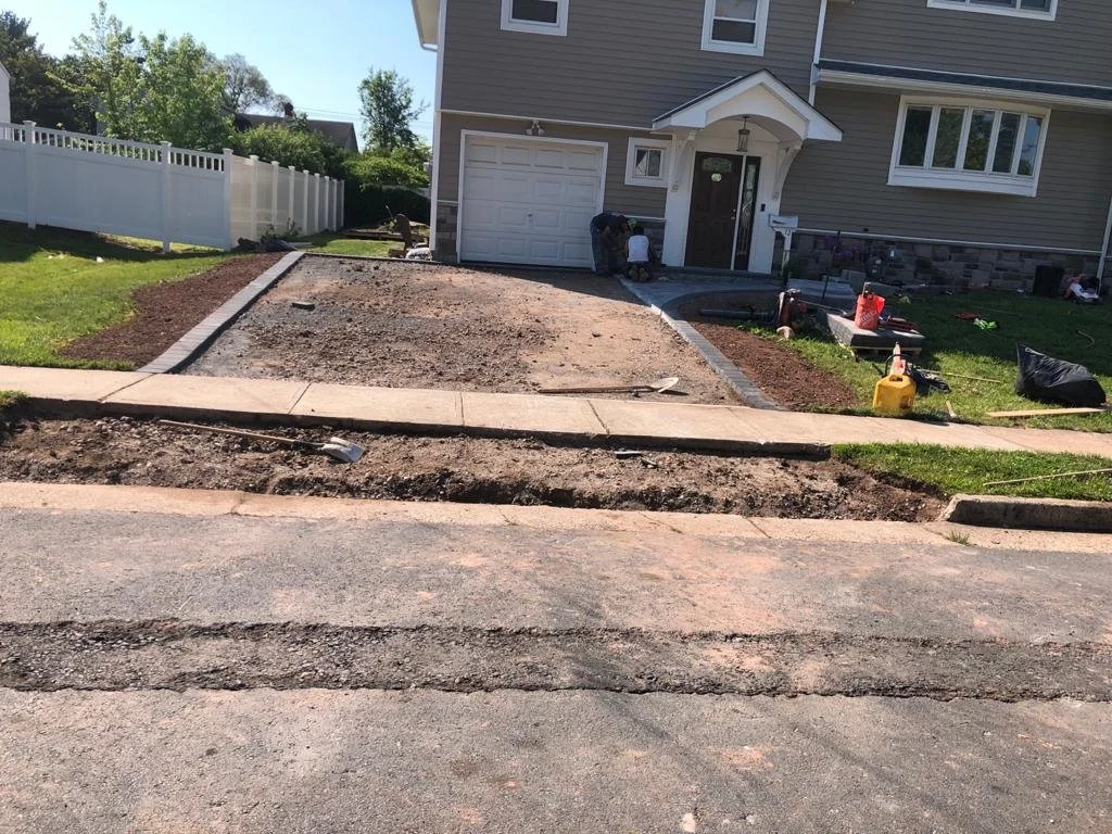 A house with a driveway under construction, with dirt and gravel where the driveway will be expanded, and construction tools and supplies nearby.