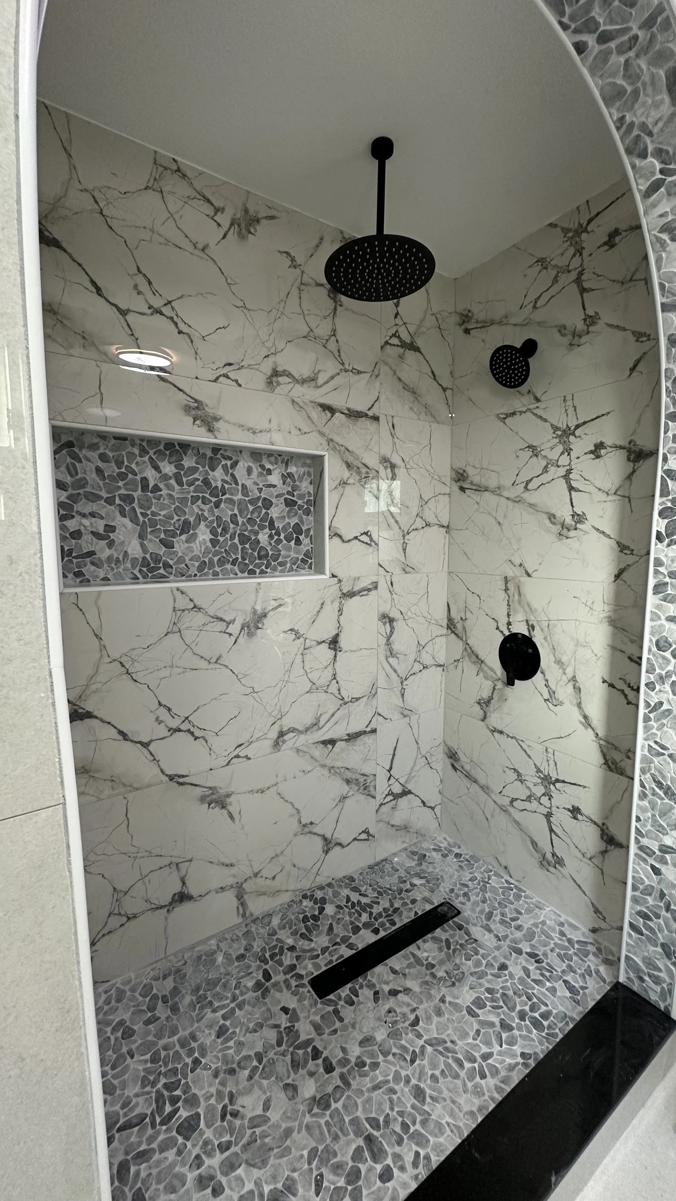 Modern shower with marble tiles, pebble stone flooring, and black fixtures.