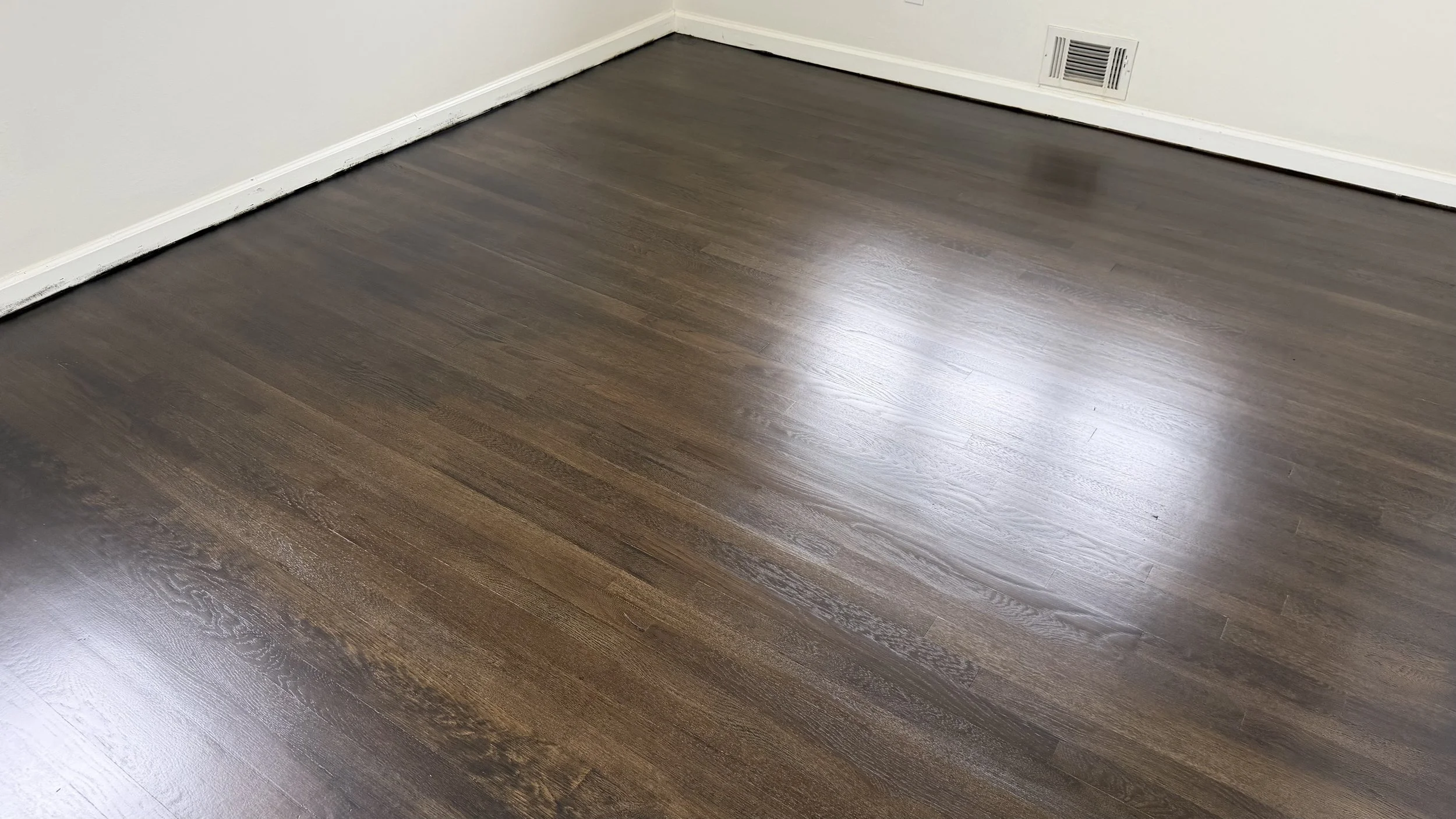 Photo of a dark brown wooden floor in a room with white walls and a vent.
