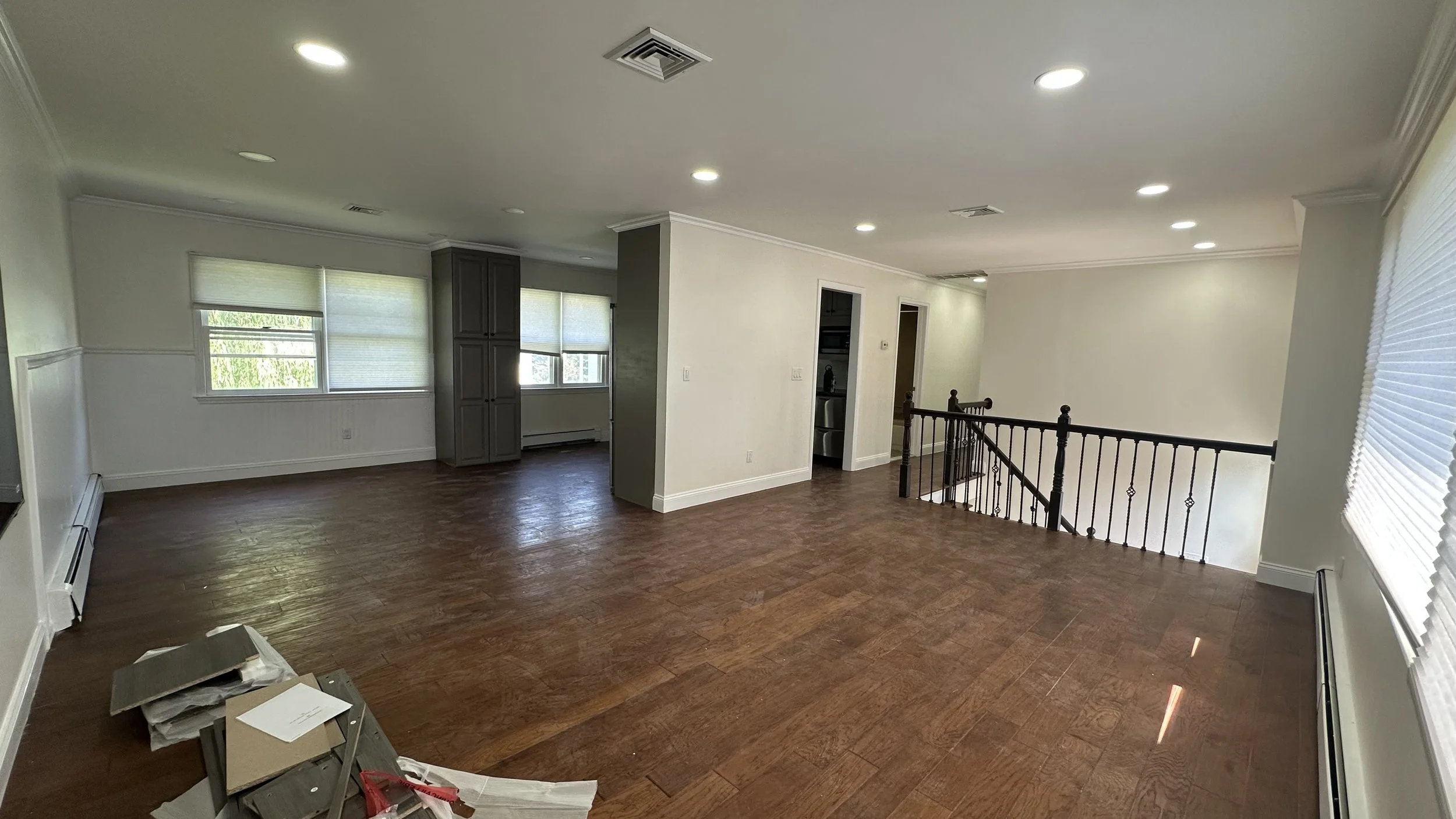 Empty living room with hardwood floors, white walls, multiple windows with blinds, and a staircase with black railing.