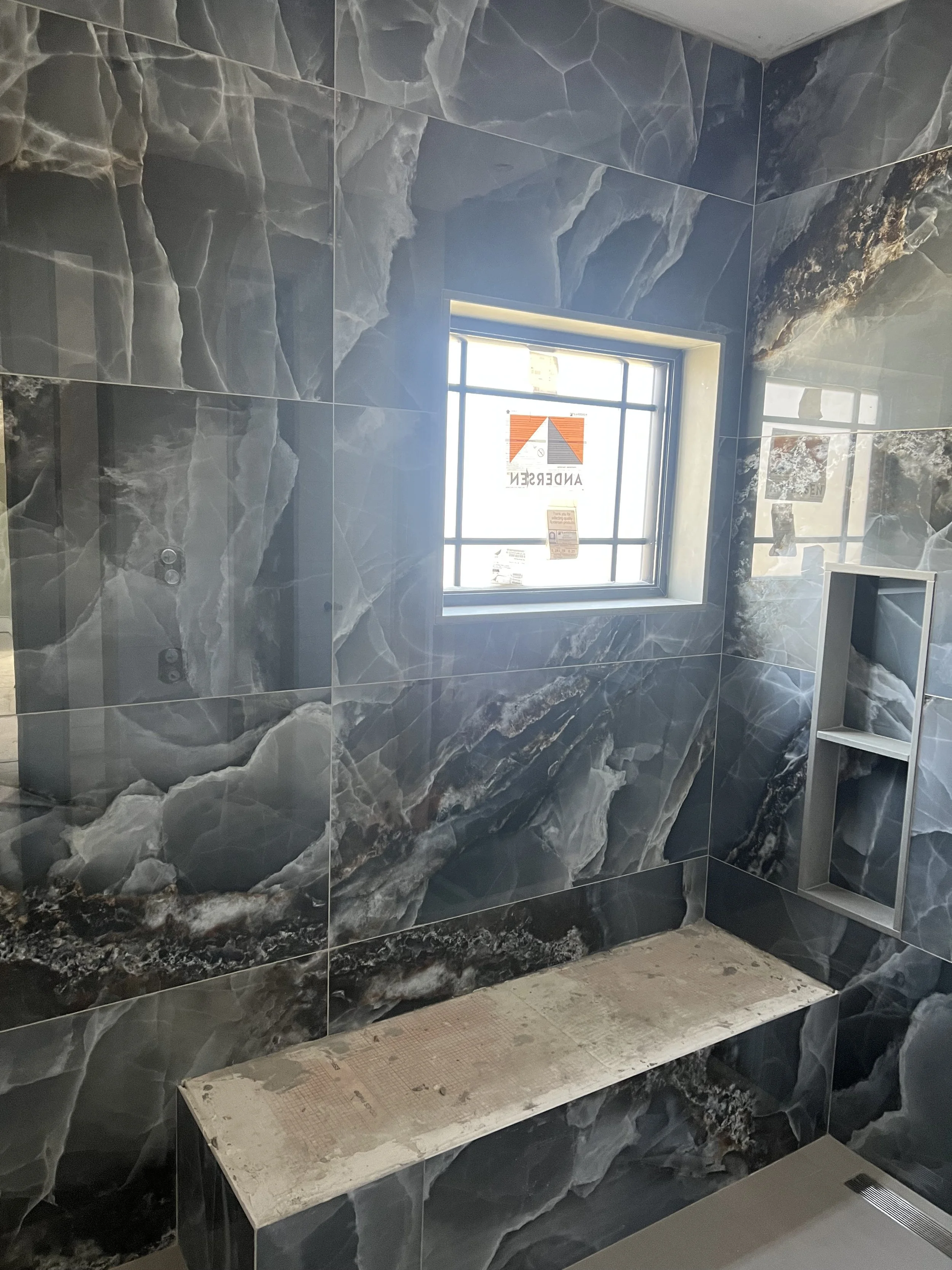Bathroom with dark, marbled tile on the walls and a large window providing natural light. There is a white, temporary bench or ledge with some construction or renovation materials underneath.
