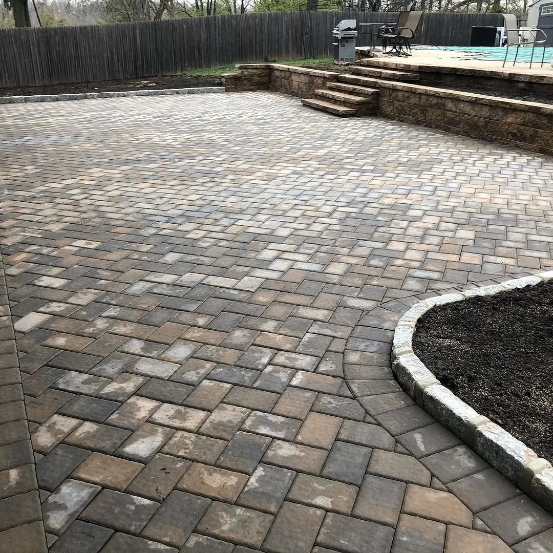 A backyard patio with interlocking brick pavers, a raised stone deck with steps, seating, and a fenced yard with trees.