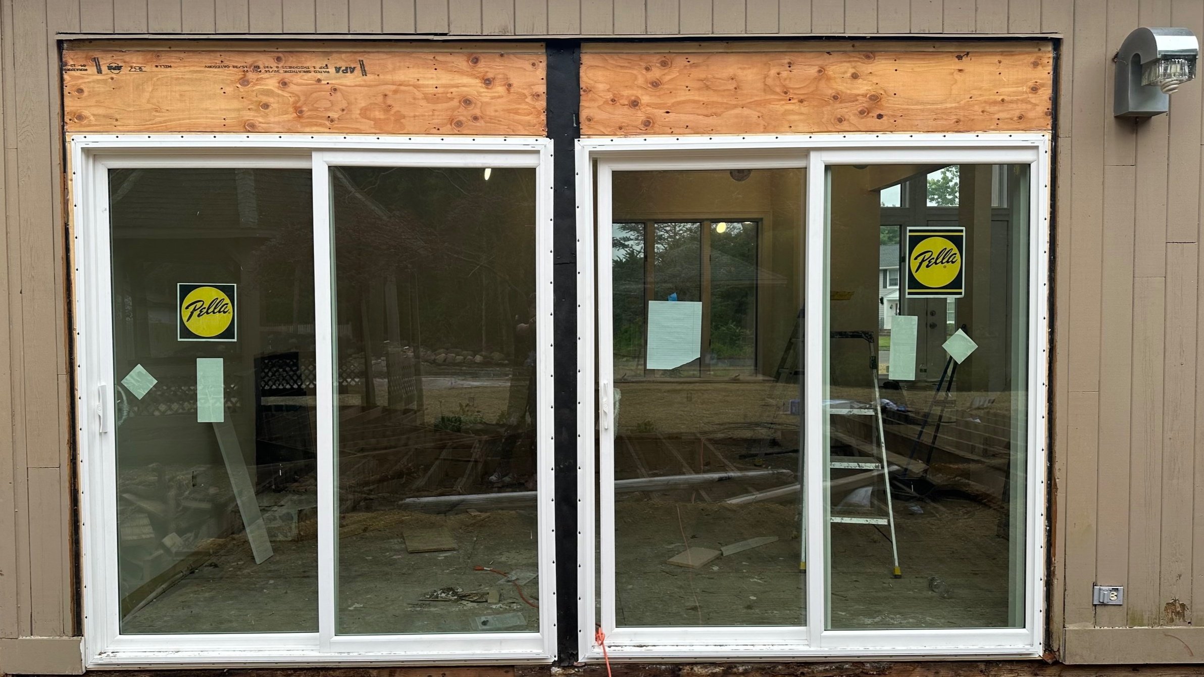 A home under construction featuring two large sliding glass doors, wooden framing sections above, interior visible through the glass with construction materials, ladder, and tools, and a yellow and black Pella window sticker.