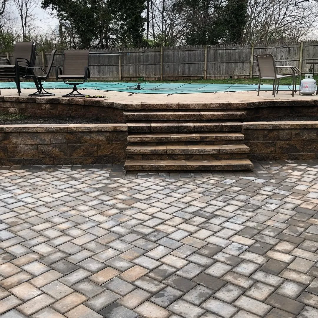 Backyard patio with stone stairs leading to a pool, surrounded by patio furniture and fencing, with some trees in the background.