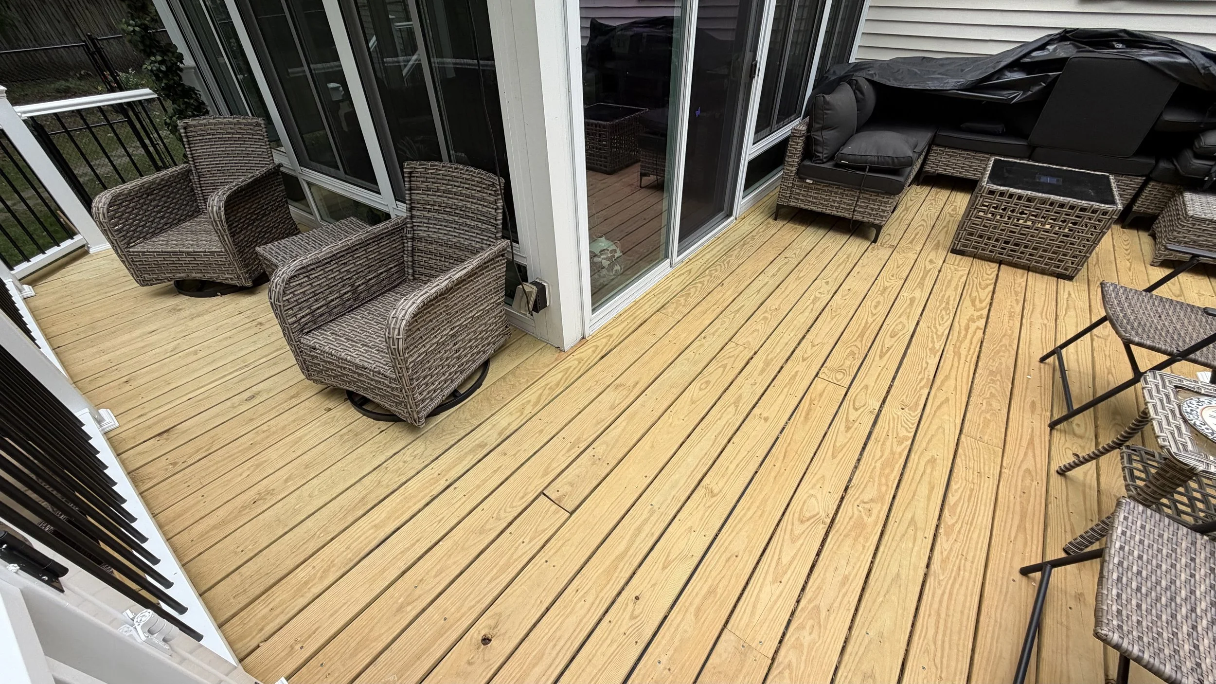 A wooden deck with outdoor furniture including four wicker chairs, a black cushioned sofa, and a table, all enclosed by a black metal railing.