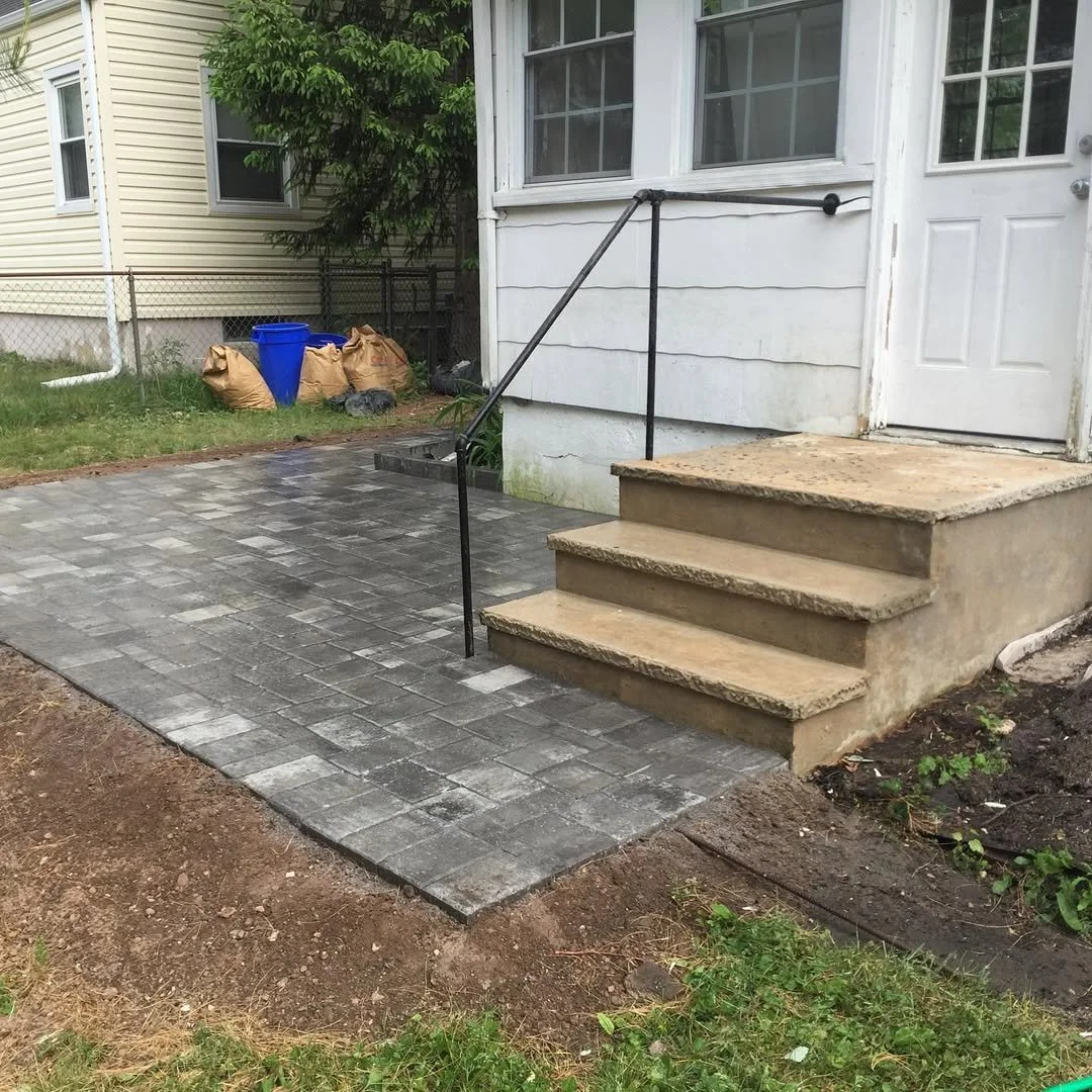 Newly installed paver walkway leading to concrete stairs with metal handrail in front of a white house door.