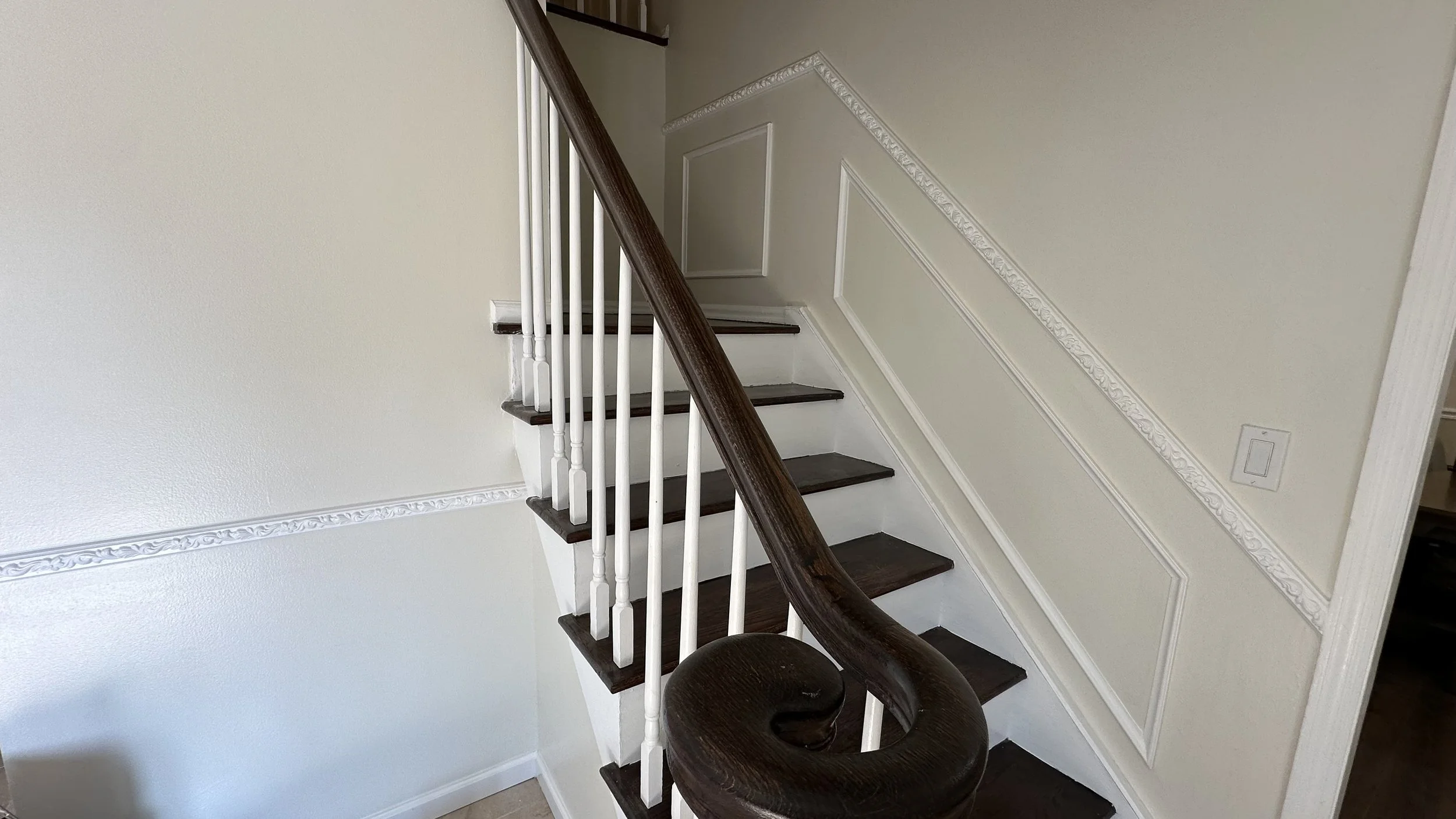 Indoor staircase with dark wooden handrail and white balusters, leading upstairs, adjacent to a cream-colored wall with decorative molding.