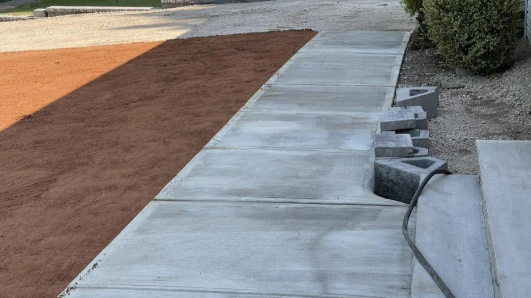 A concrete sidewalk with a freshly poured section, adjacent to a dirt area and some construction supplies like cinder blocks.
