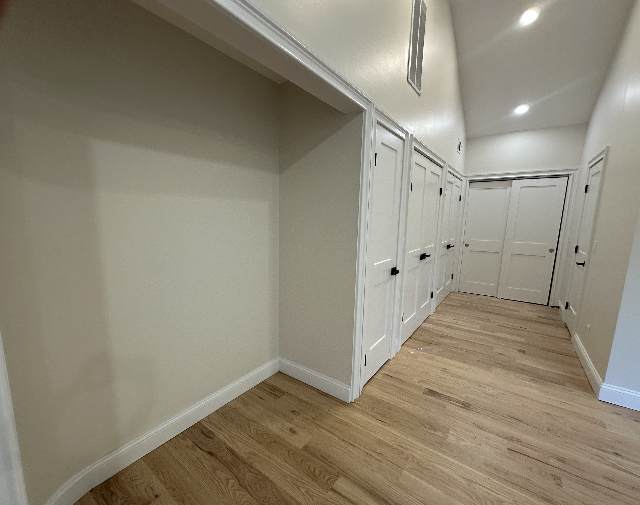 Empty hallway with beige walls, wooden floor, white closet doors with black handles, and recessed ceiling lights.