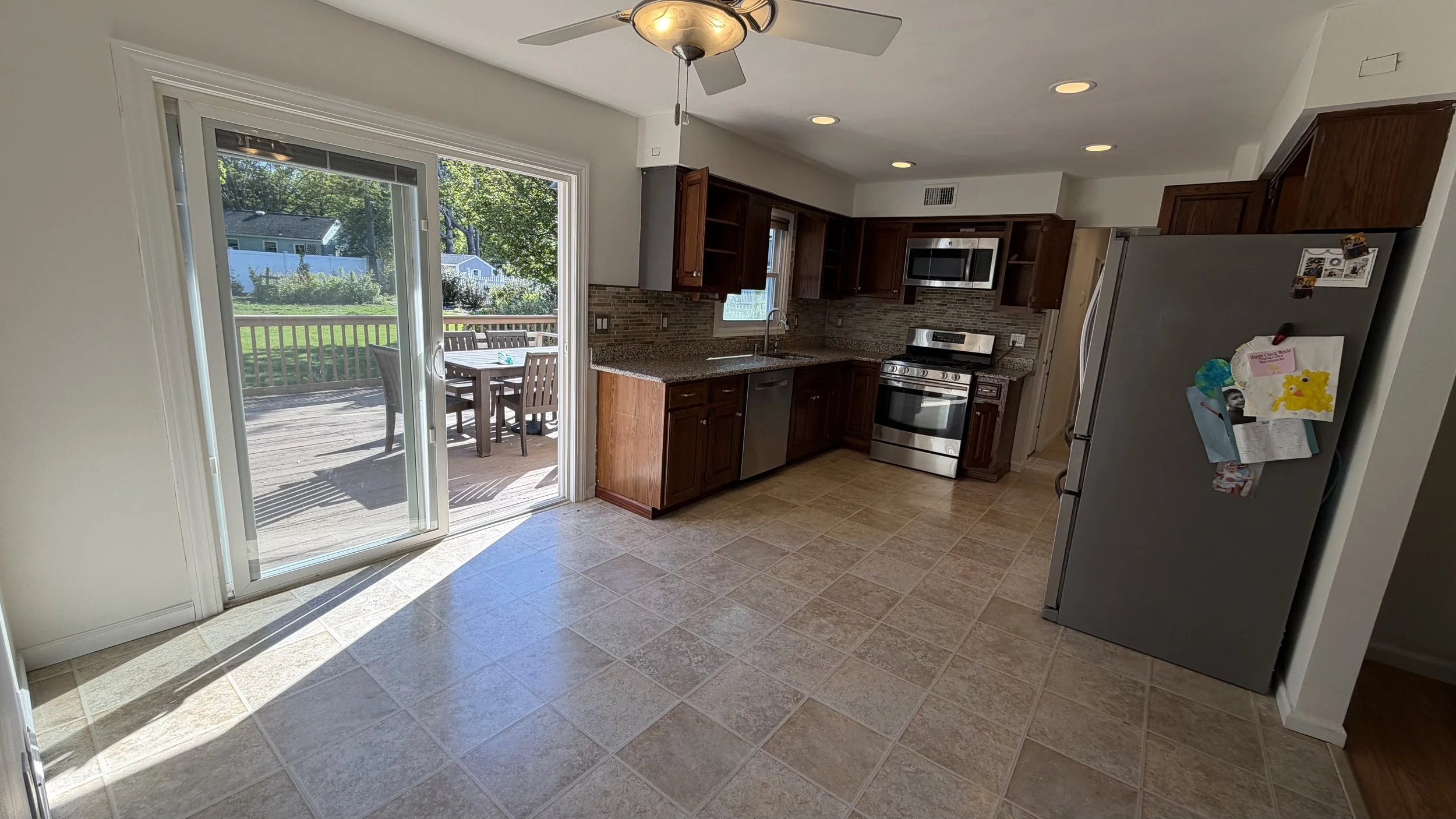 A kitchen with wooden cabinets, stainless steel appliances, and a sliding glass door leading to an outdoor deck with patio furniture. The room has beige tiled flooring, a ceiling fan, and recessed lighting.