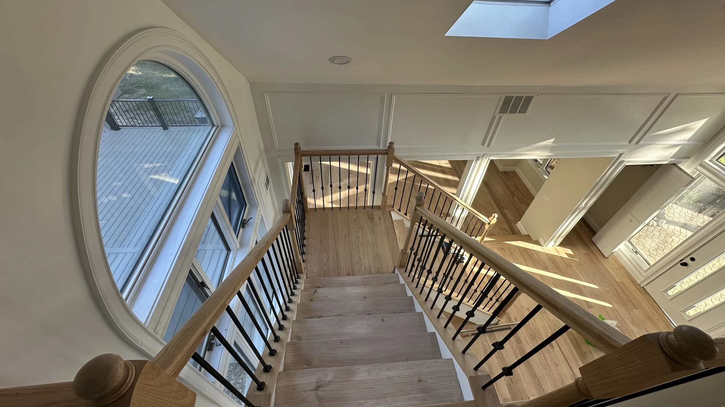 A view of a staircase from the top, showing wooden steps with black metal balusters, leading down to a hallway with hardwood floors. Sunlight streams through large windows, illuminating the space.