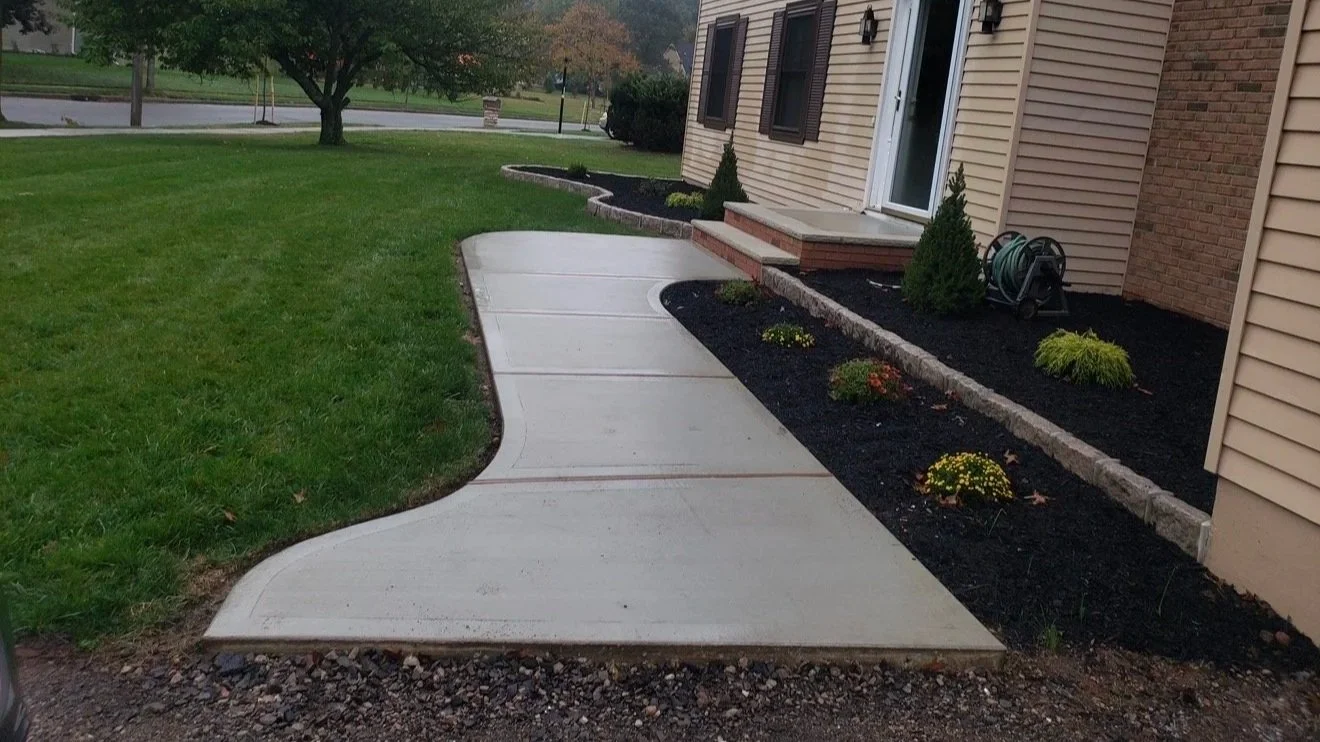 Newly poured concrete sidewalk with decorative curved edges in front of a house, with a garden bed on the side containing small shrubs and flowers, and a grassy lawn nearby.
