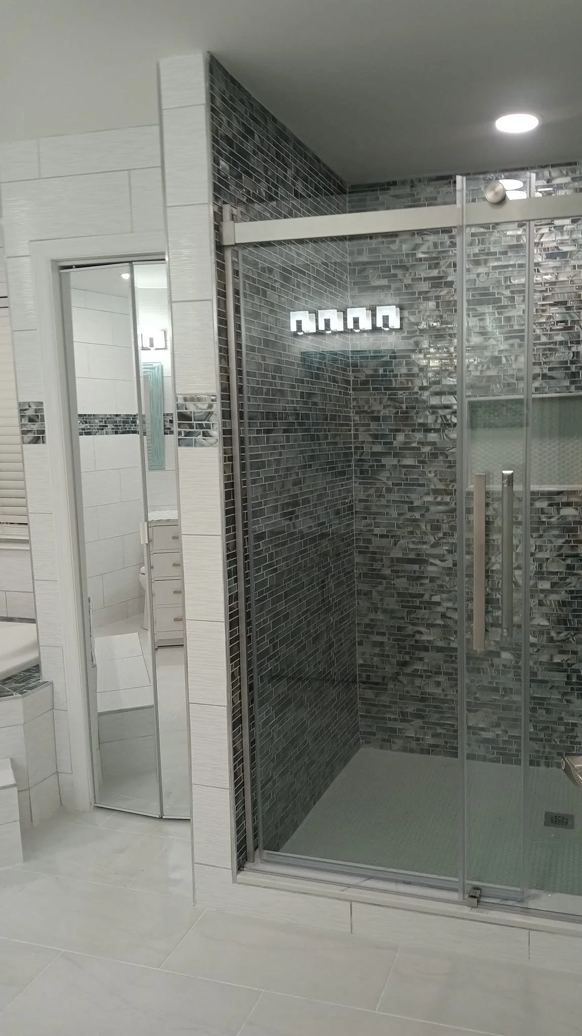 Bathroom shower with glass enclosure, dark gray tile interior, and a white tiled wall with a small mirror and a towel rack.