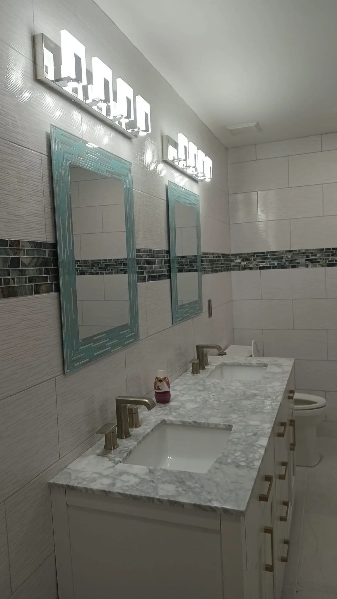 Bathroom with two mirrors, marble countertop with sinks, modern faucets, tile walls, and a toothbrush holder.