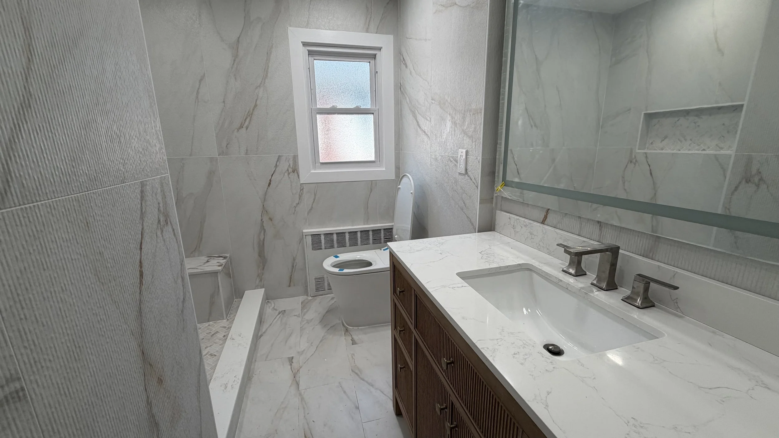 Modern bathroom with marble walls, a window, a toilet, and a wooden vanity with a white marble countertop and an integrated sink.