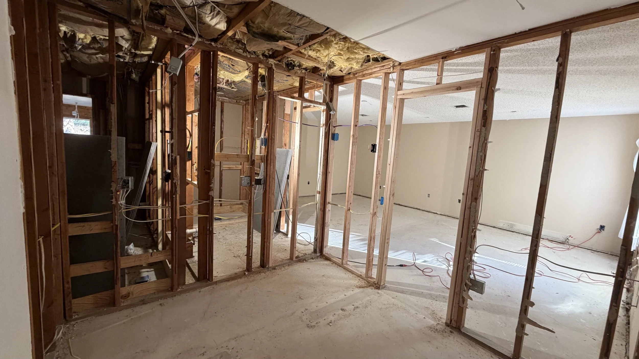 Interior of a house under renovation with exposed wooden wall studs, electrical wiring, and insulation at the ceiling, with the floor unfinished and debris visible.