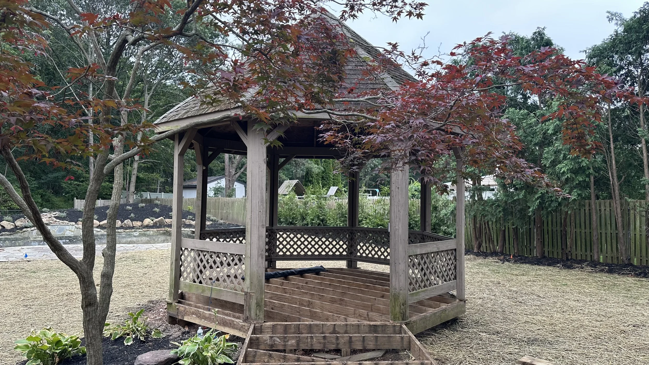 A wooden gazebo with lattice sides and a shingled roof, surrounded by trees and a backyard with a fence and a pond in the background.