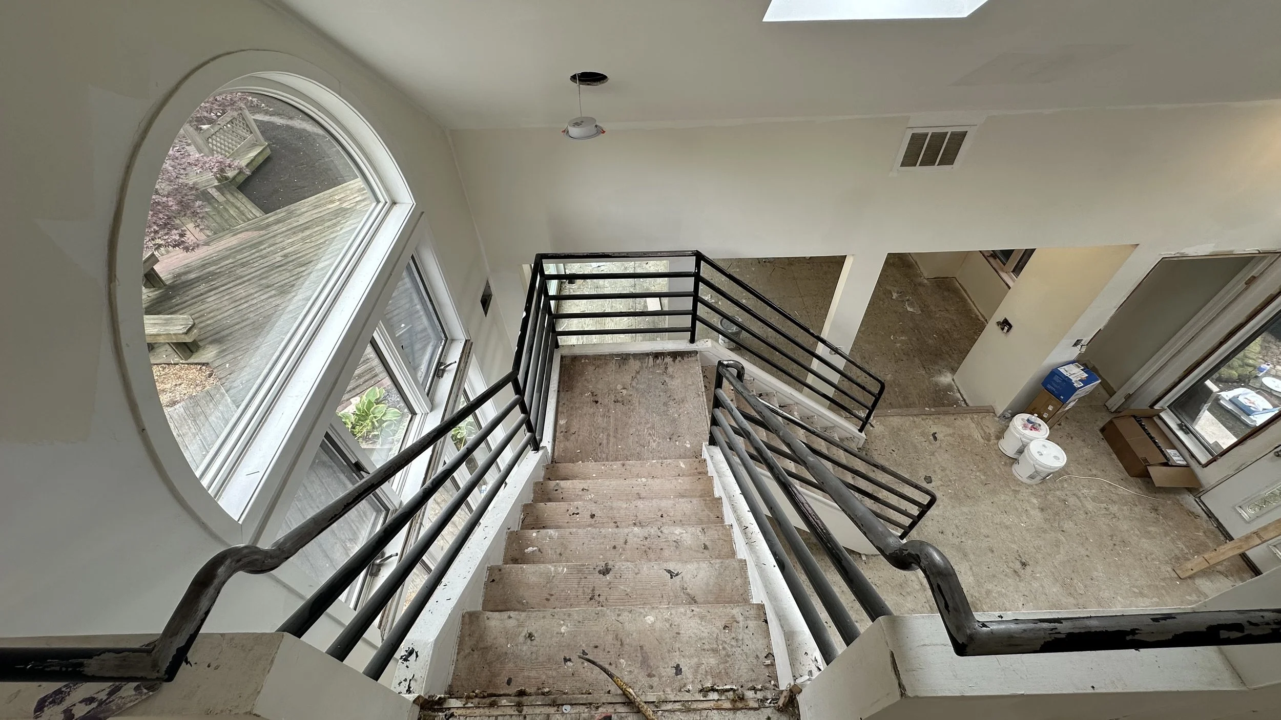 Interior of a house under renovation, viewed from the top of a staircase with black metal handrails, showing bare wooden stairs, a large arched window overlooking a deck, and construction supplies on the floor.
