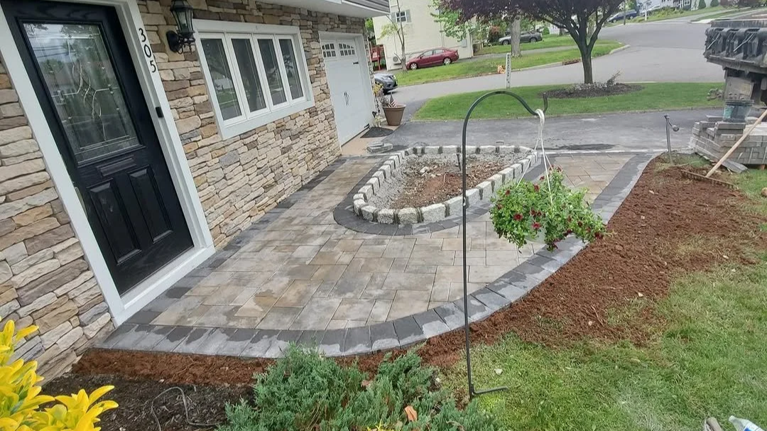 The front yard of a house under renovation with a newly paved pathway, a raised flower bed, and a hanging flower basket.